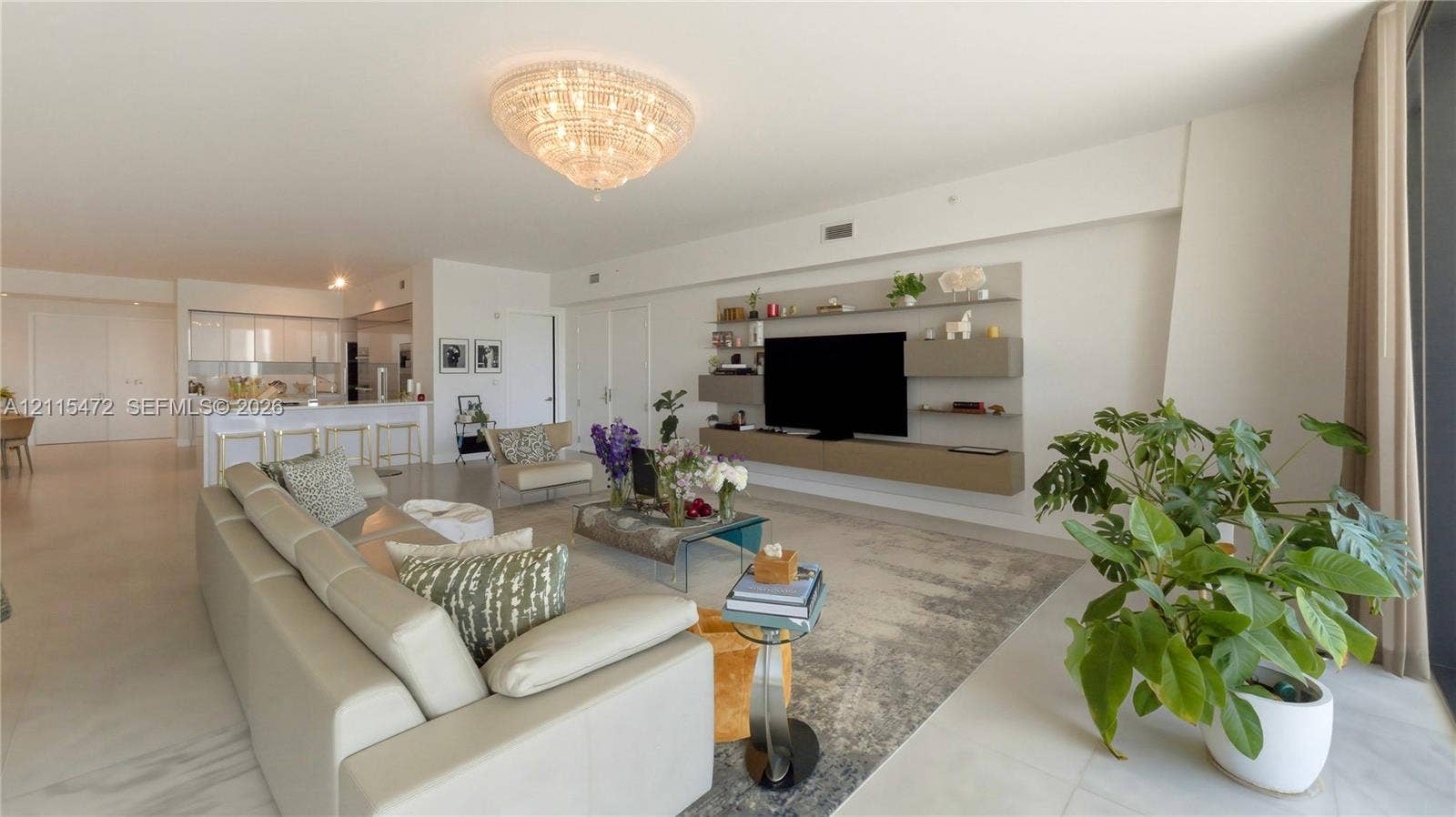 Open-concept living space featuring large-format tile flooring, a statement crystal chandelier, custom wall-mounted floating cabinetry, and a kitchen island with stone-finish countertops