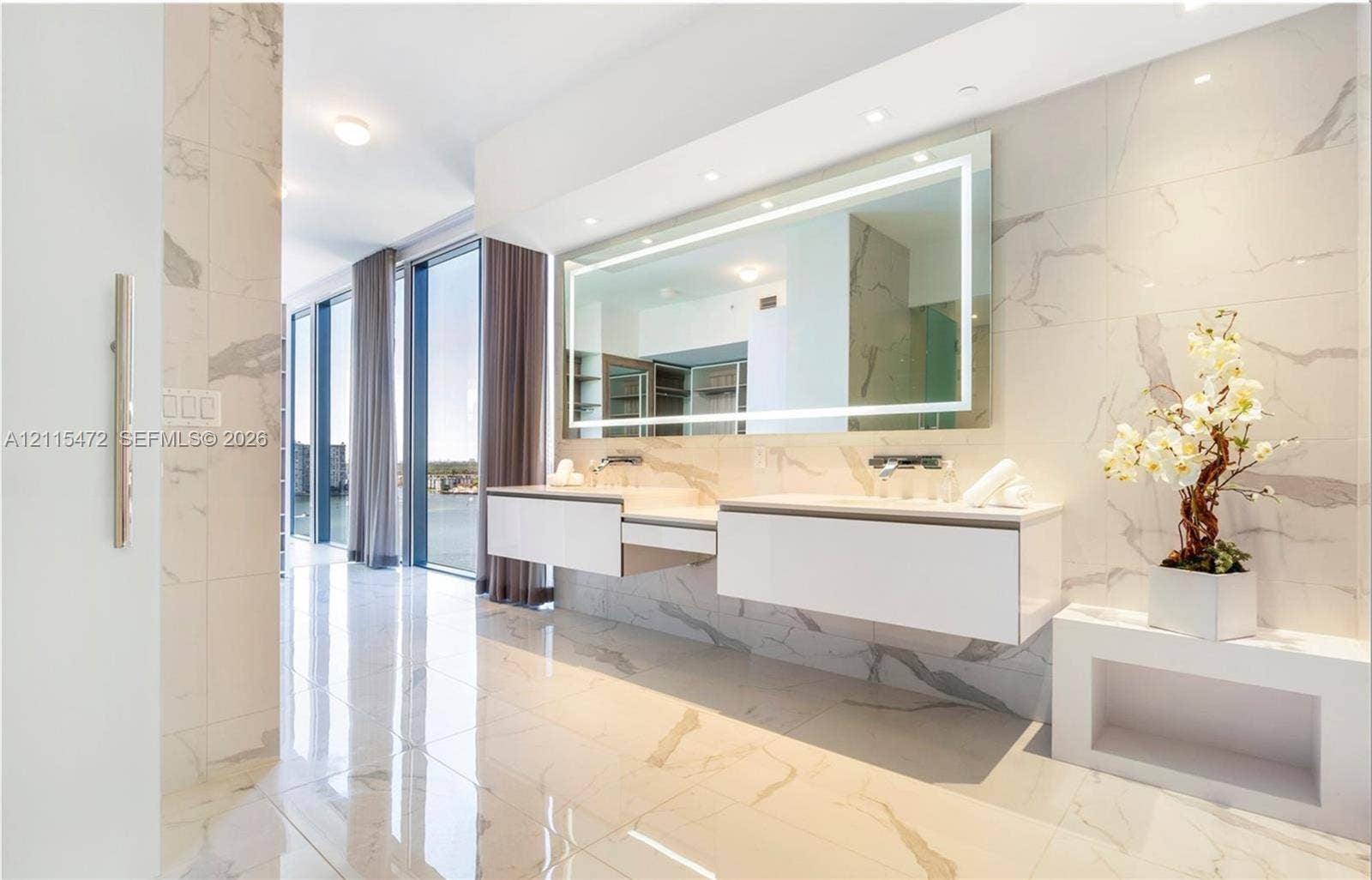 Floating dual vanity with integrated sinks, large LED-backlit mirror, floor-to-ceiling stone-look tiling, and expansive floor-to-ceiling windows