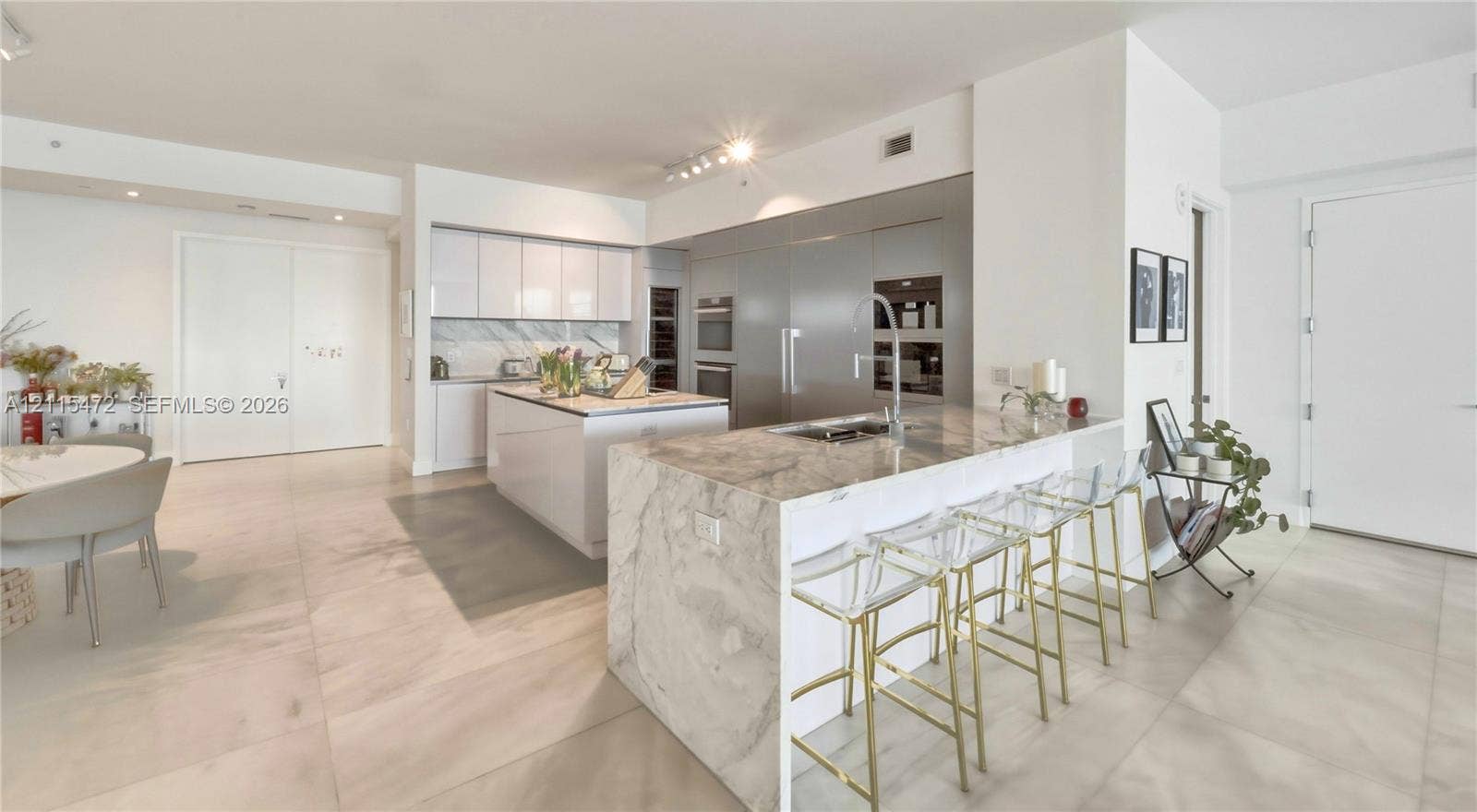 Modern kitchen featuring an expansive waterfall-edge island, stone countertops, integrated stainless steel appliances, flat-panel cabinetry, and large-format tile flooring