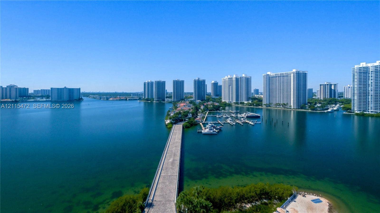 Aerial perspective of a coastal waterway featuring a paved causeway, deep-water access, a private marina, and surrounding high-rise structures