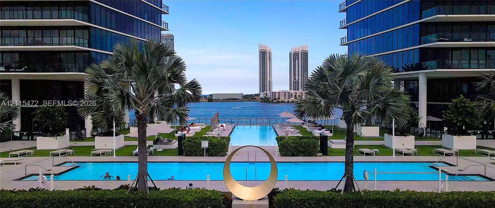 Waterfront infinity-edge swimming pool featuring mature palm trees, manicured landscaping, paved deck, and sculptural metal art