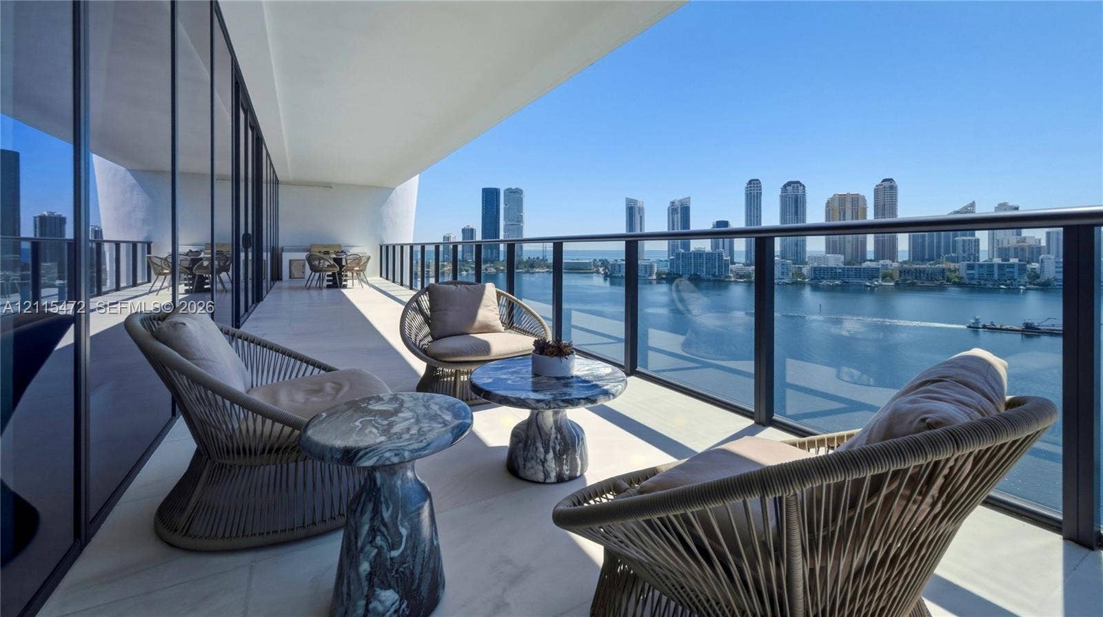 Expansive covered balcony featuring white tile flooring, floor-to-ceiling glass wall, and a full-perimeter metal railing overlooking the water and urban skyline