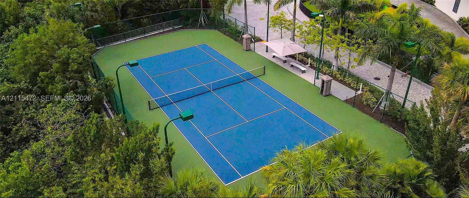 Private hard-court tennis facility featuring perimeter fencing, integrated lighting, paved pedestrian walkway, and mature landscaping