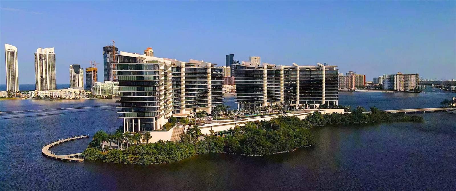 Waterfront high-rise complex featuring expansive glass-walled balconies, ground-level swimming pools, and a private pedestrian pier on a coastal peninsula