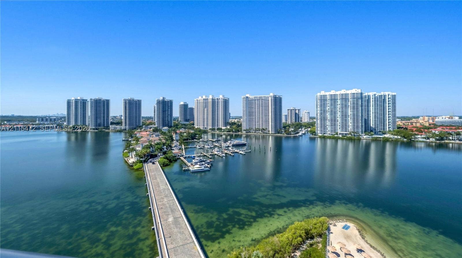 Elevated aerial perspective featuring panoramic water expanses, a private marina with multiple slips, a concrete causeway, and expansive coastal high-rise scenery