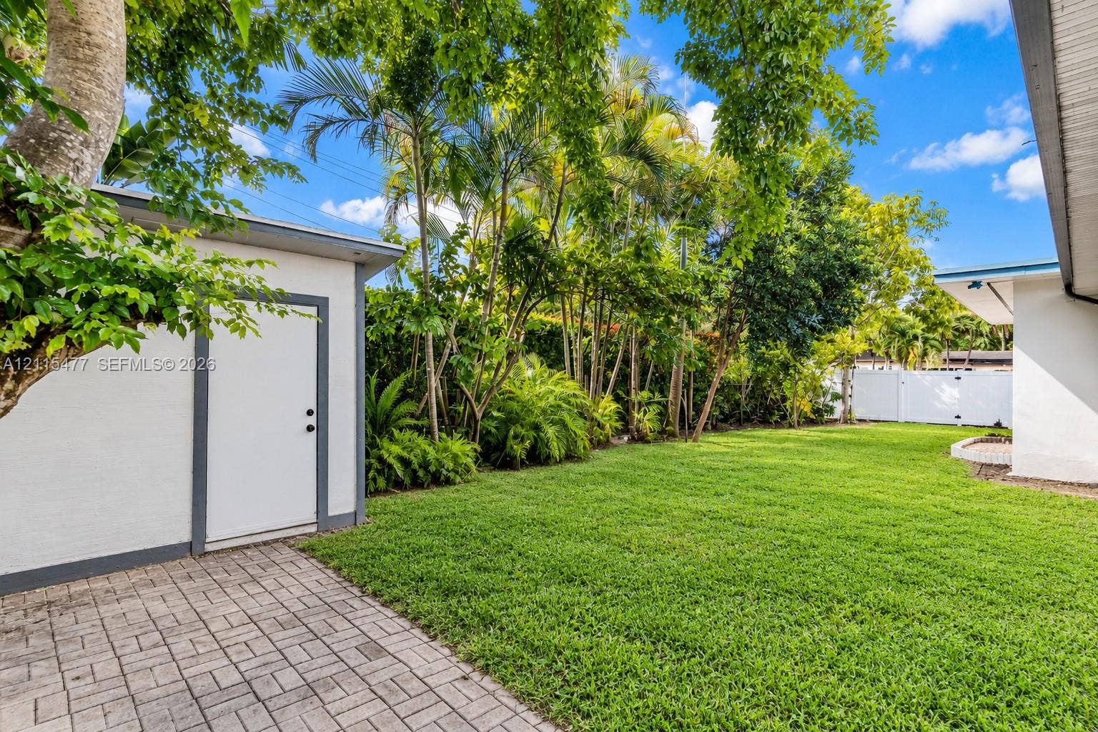 2510 NE 208th Terrace, Miami, FL 33180 Backyard featuring a paved stone patio, detached storage structure, manicured lawn, and mature perimeter landscaping enclosed by white fencing