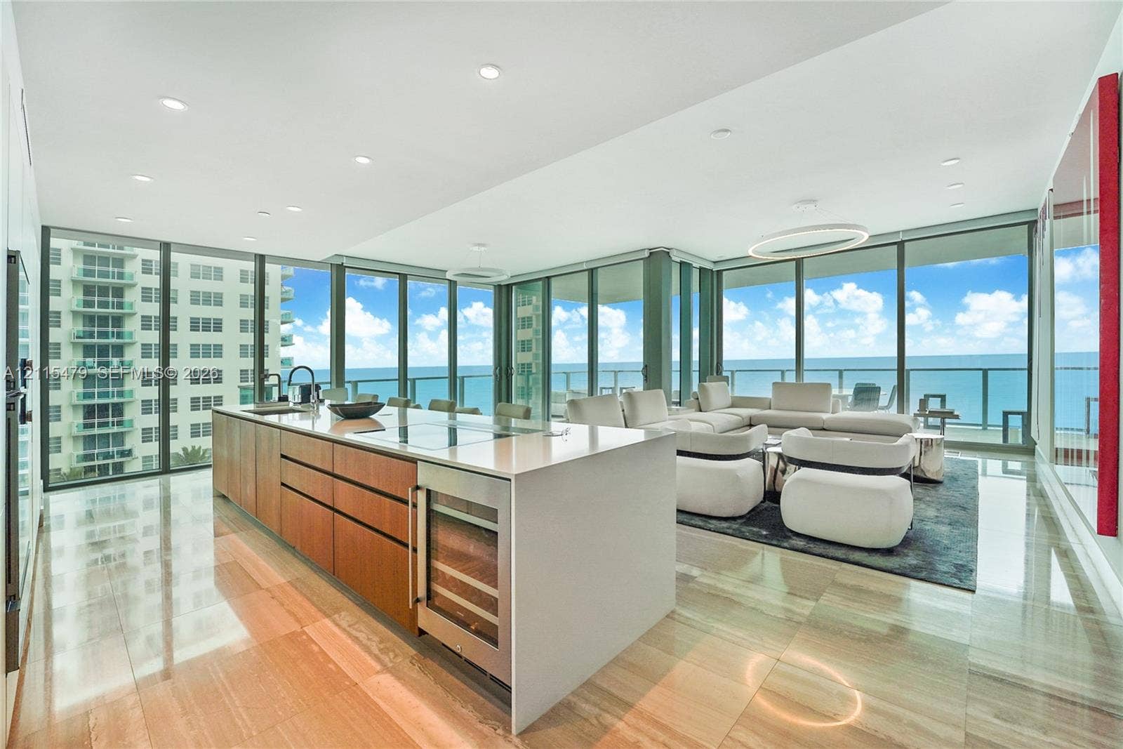Open-concept kitchen featuring an expansive quartz-topped island, integrated wine refrigeration, warm-tone wood cabinetry, recessed ceiling lighting, and floor-to-ceiling glass panel walls