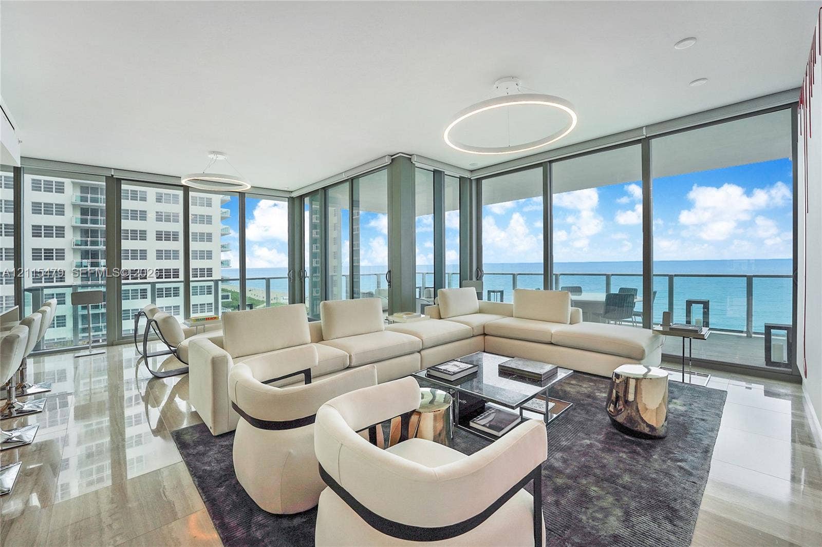Floor-to-ceiling glass windows, polished tile flooring, circular pendant lighting, and an expansive balcony featuring a glass railing and ocean scenery