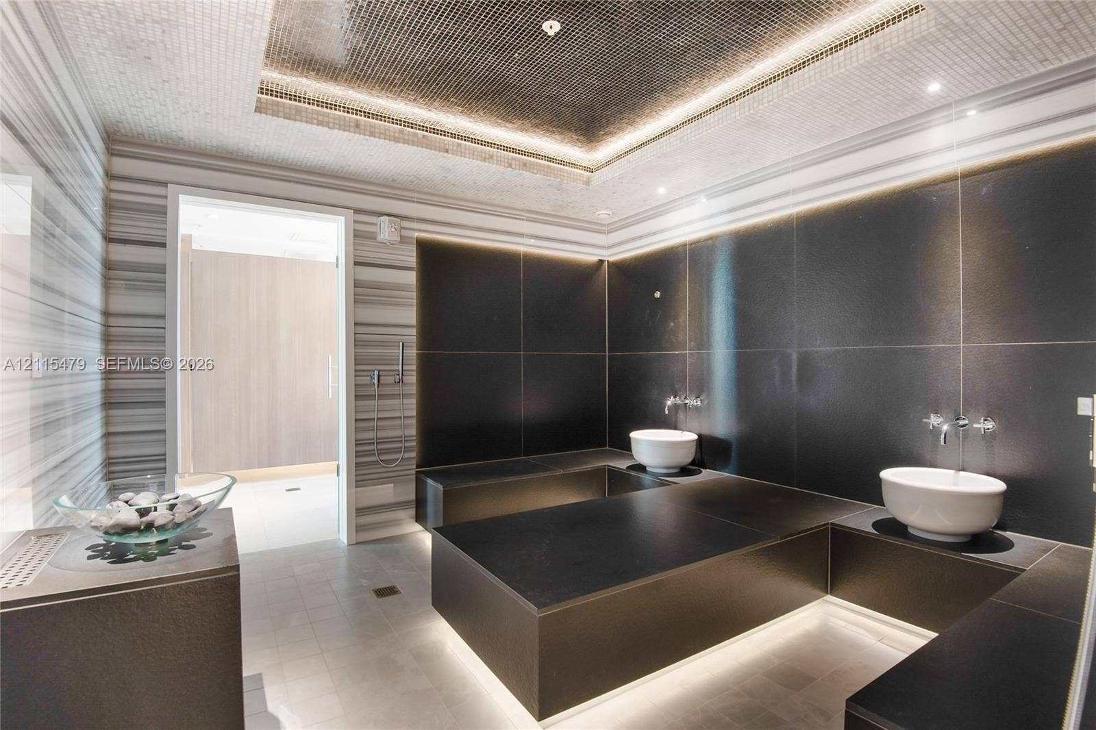 Custom bathroom featuring floor-to-ceiling linear-patterned tile, expansive black-tiled platform surfaces, dual vessel sinks with wall-mounted faucets, and a recessed tray ceiling with integrated perimeter lighting