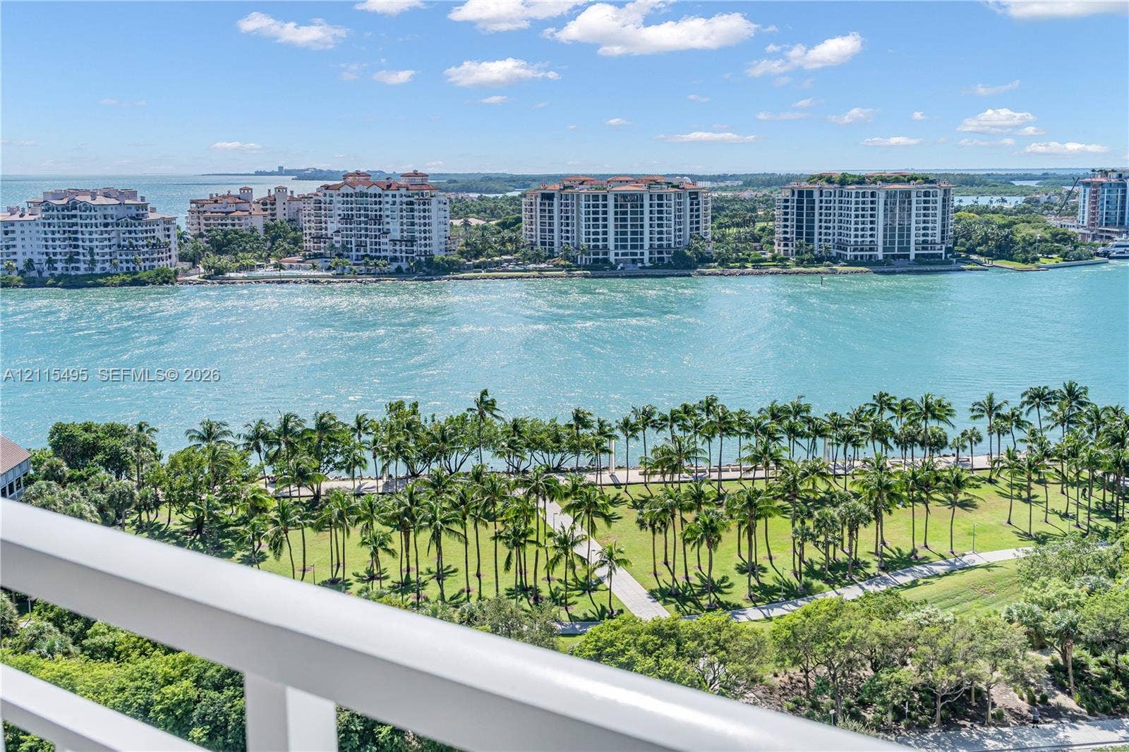 Elevated balcony with a white metal railing, showcasing coastal scenery, waterfront expanse, and a landscaped park with mature palm trees