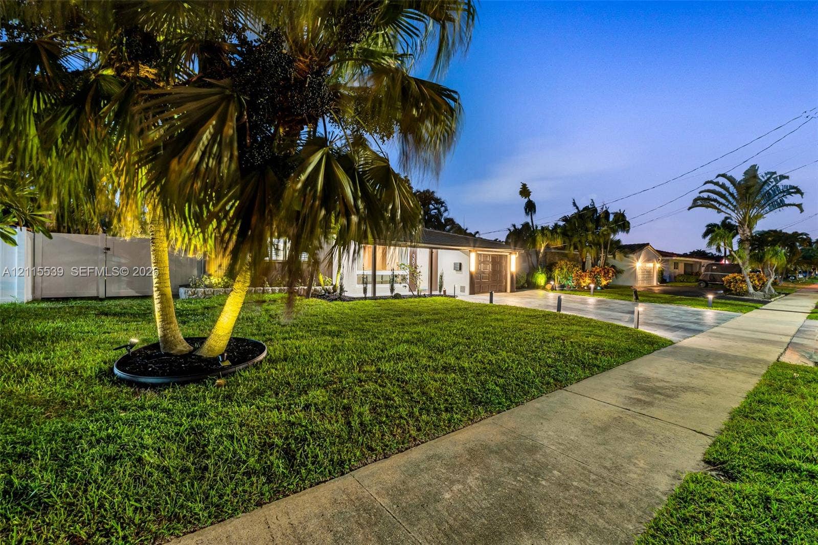381 SE 6th Terrace, Pompano Beach, FL 33060 Exterior facade featuring a concrete driveway, manicured lawn, mature palm trees, white vinyl privacy fencing, and exterior landscape lighting