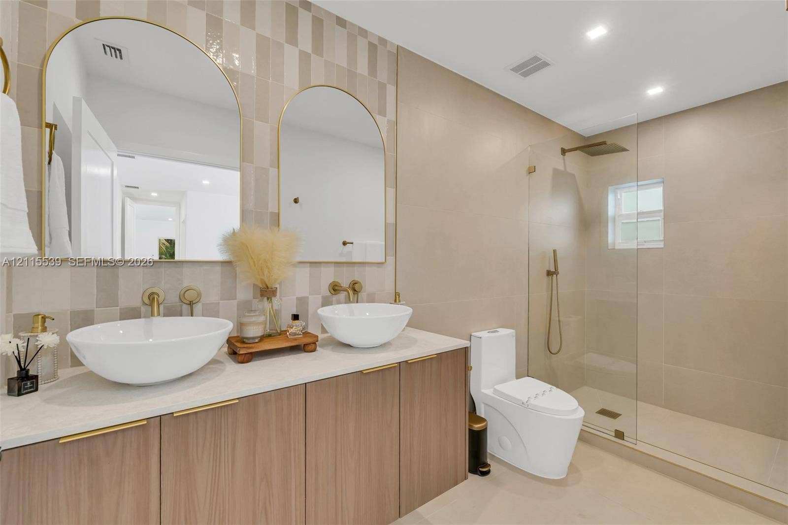 381 SE 6th Terrace, Pompano Beach, FL 33060 Dual vanity featuring vessel sinks, quartz countertops, wood-finish cabinetry, arched mirrors with gold-toned frames, and a walk-in shower with a glass enclosure