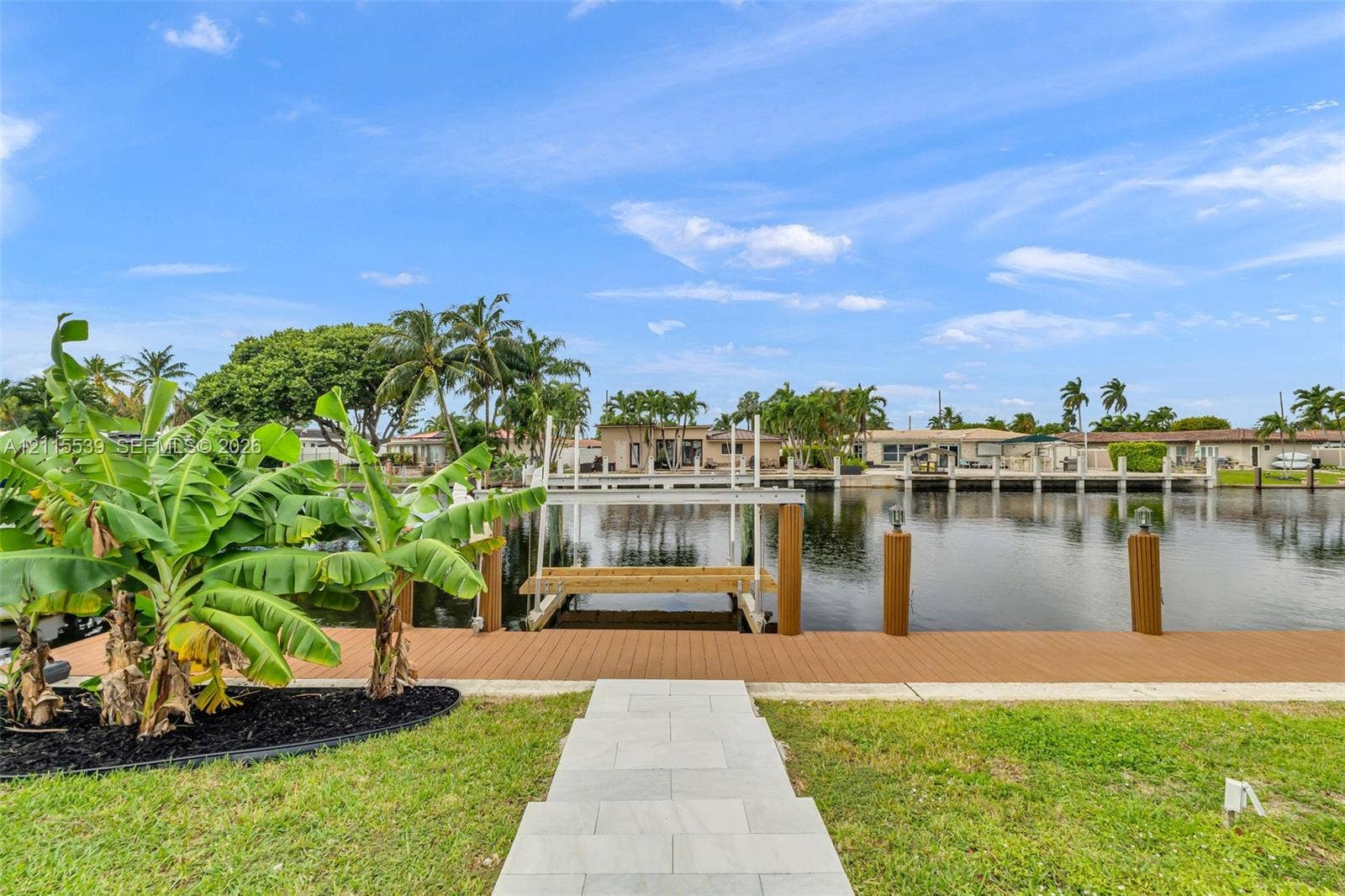 381 SE 6th Terrace, Pompano Beach, FL 33060 Waterfront property featuring a composite deck, private boat lift, and anchored wooden pilings, complemented by a paver stone walkway and tropical landscaping