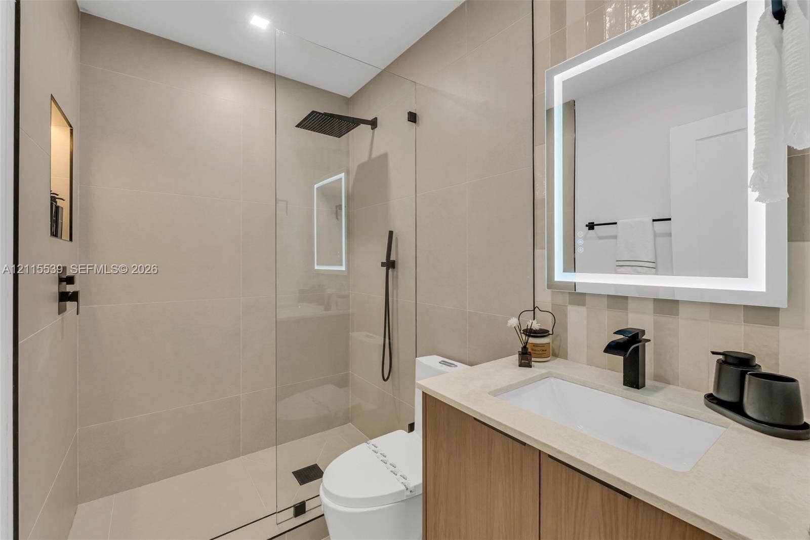 381 SE 6th Terrace, Pompano Beach, FL 33060 Bathroom featuring a wood-finish vanity with a light-toned stone countertop, a glass-enclosed walk-in shower with matte black fixtures, a built-in shower niche, and an integrated LED-backlit mirror
