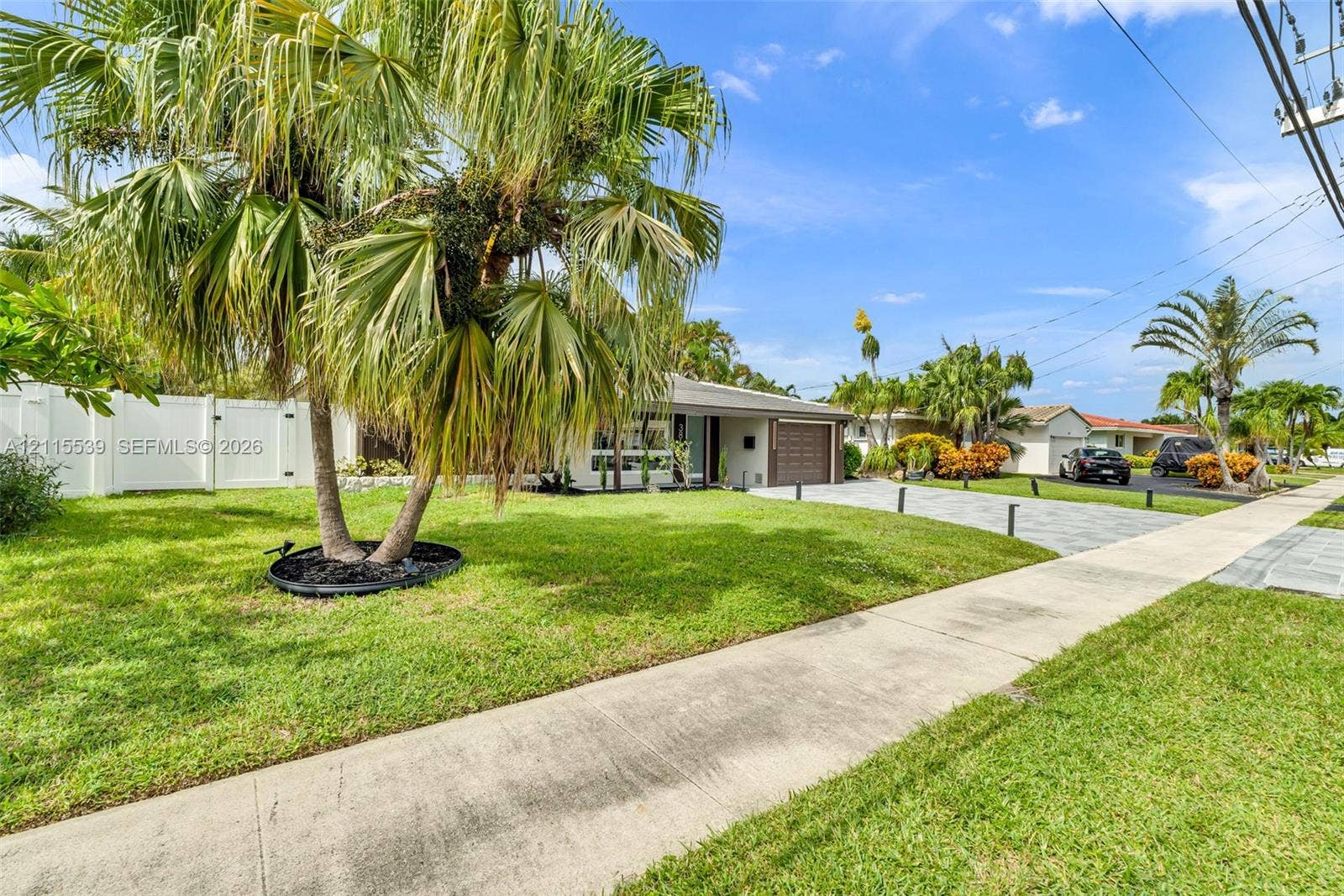 381 SE 6th Terrace, Pompano Beach, FL 33060 Single-story residence featuring a paver driveway, an attached garage, white privacy fencing, and established palm trees