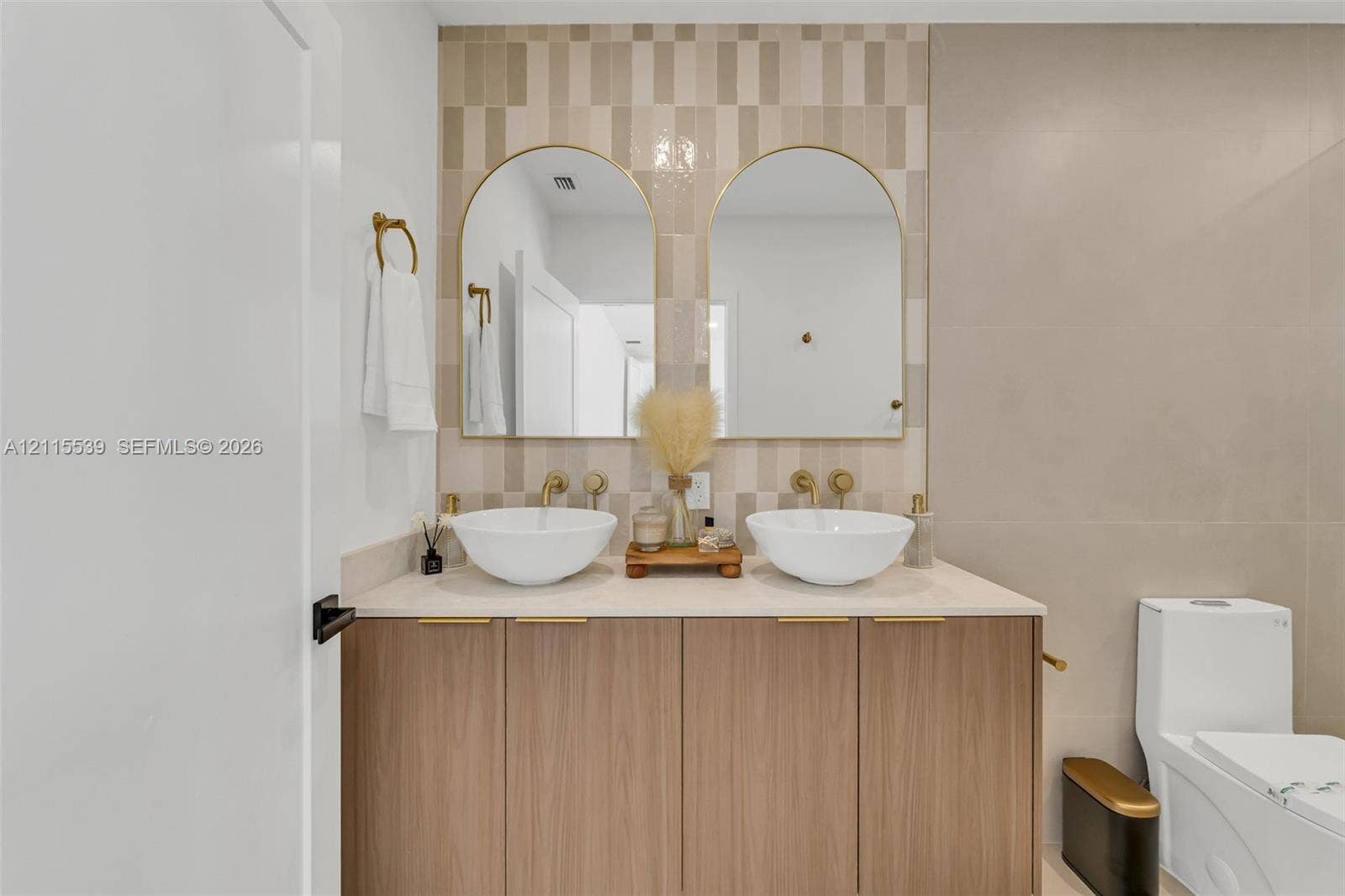 381 SE 6th Terrace, Pompano Beach, FL 33060 Double vanity featuring wood-finish cabinetry, white vessel sinks, gold-tone wall-mounted faucets, arched mirrors, and a patterned tile backsplash