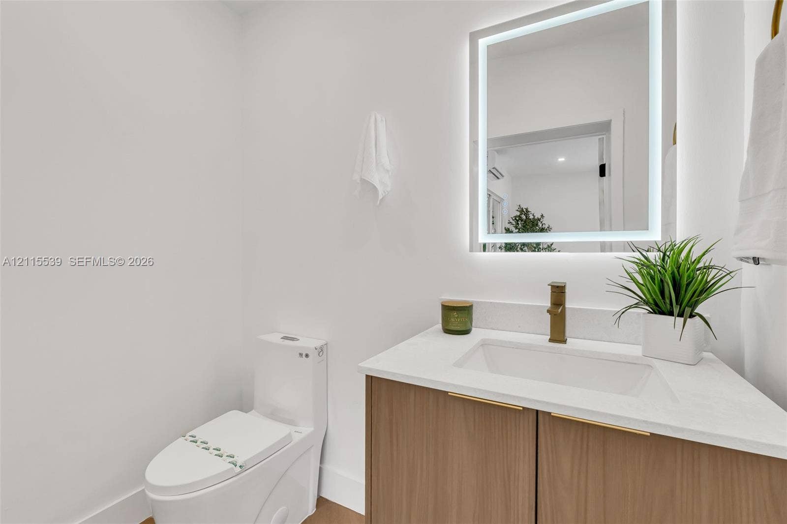 381 SE 6th Terrace, Pompano Beach, FL 33060 Wood-finish vanity cabinet, white solid-surface countertop, rectangular undermount sink, brass-tone faucet, and rectangular LED-backlit mirror