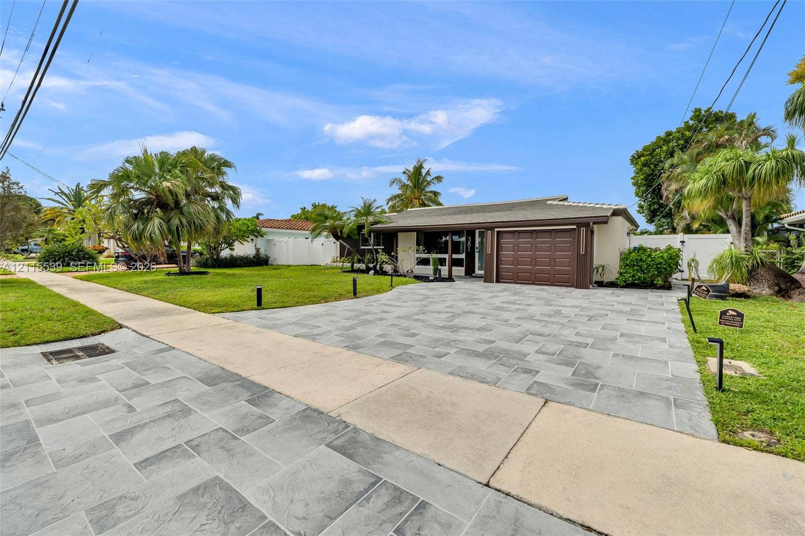 381 SE 6th Terrace, Pompano Beach, FL 33060 Single-story residence featuring a stone-paver driveway, attached garage with a multi-panel door, and white vinyl perimeter fencing
