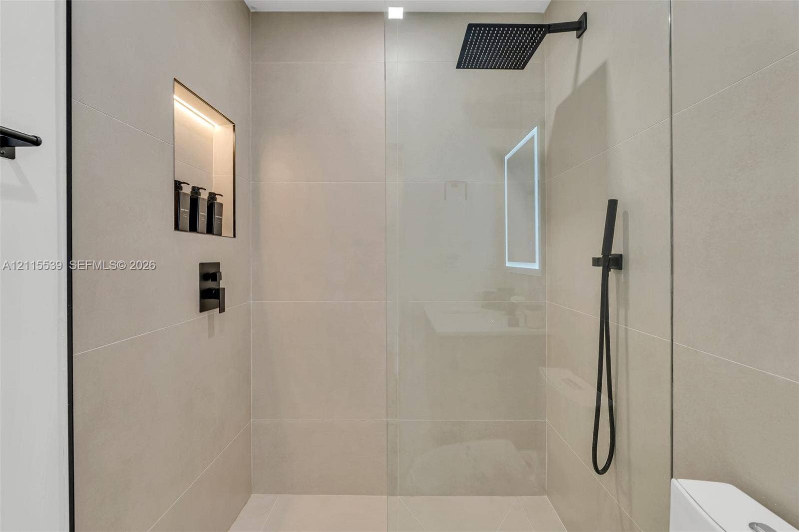 381 SE 6th Terrace, Pompano Beach, FL 33060 Walk-in shower featuring large-format neutral tile, frameless glass enclosure, matte black rainfall showerhead, integrated handheld sprayer, and recessed tiled niche with lighting