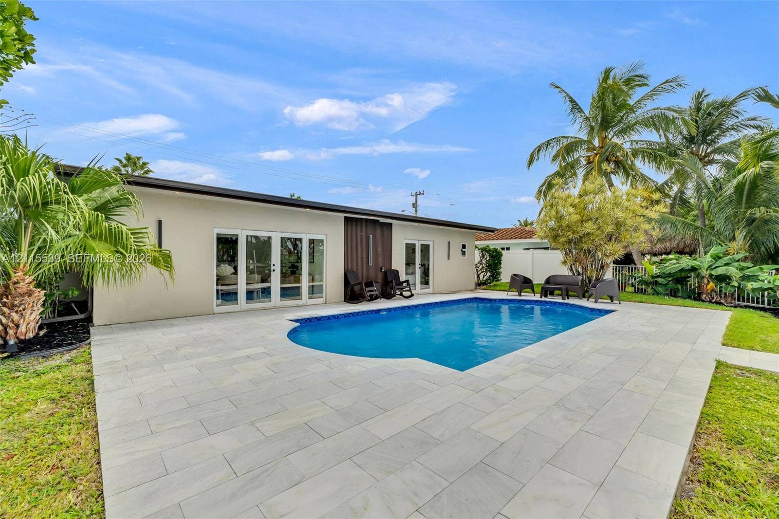 381 SE 6th Terrace, Pompano Beach, FL 33060 Rear exterior featuring an in-ground swimming pool, expansive stone paver patio, multi-panel glass door systems, and mature landscaping