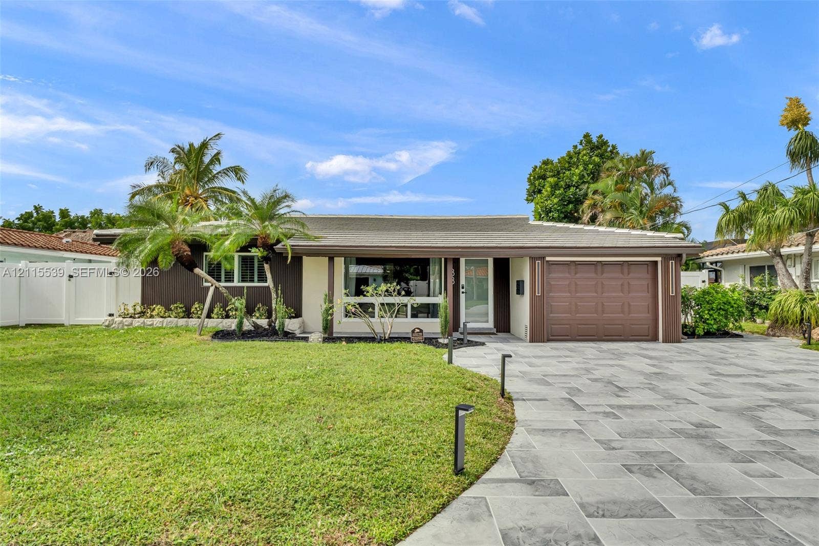 381 SE 6th Terrace, Pompano Beach, FL 33060 Single-story exterior featuring a stone-paver driveway, attached garage, large picture window, and landscaped lawn with palm trees