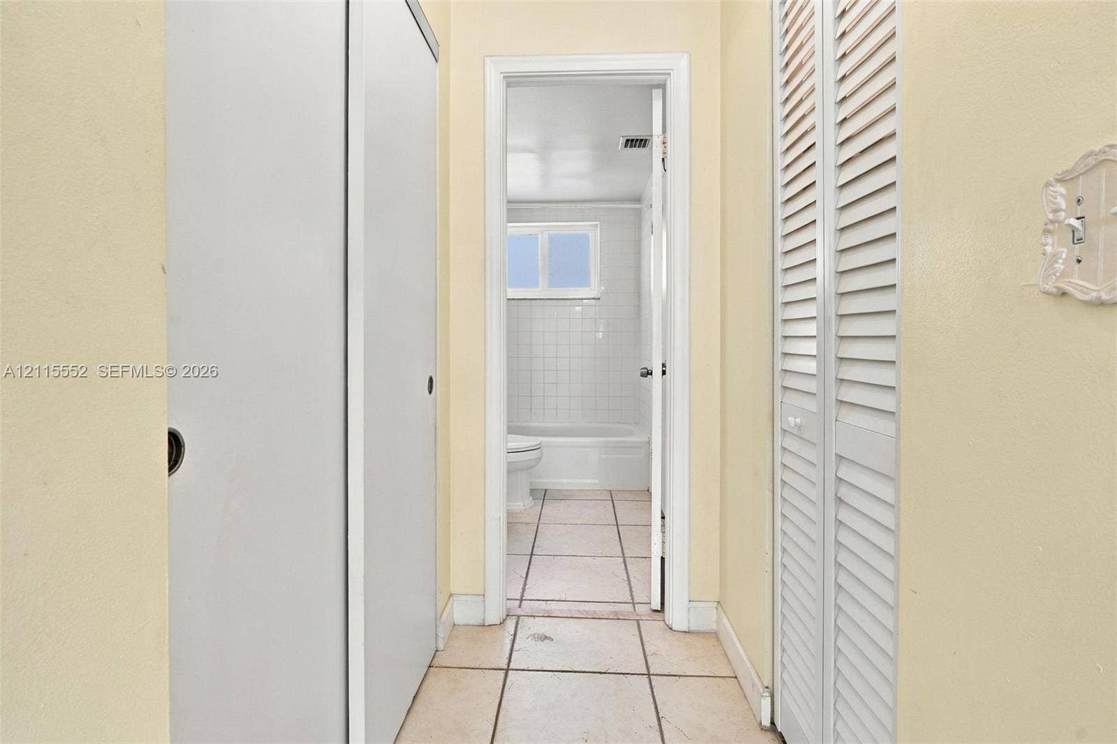 7805 Camino Real, #H-220, Miami, FL 33143 Hallway featuring dual sets of white louvered closet doors, tile flooring, and a doorway revealing a tiled tub surround and window