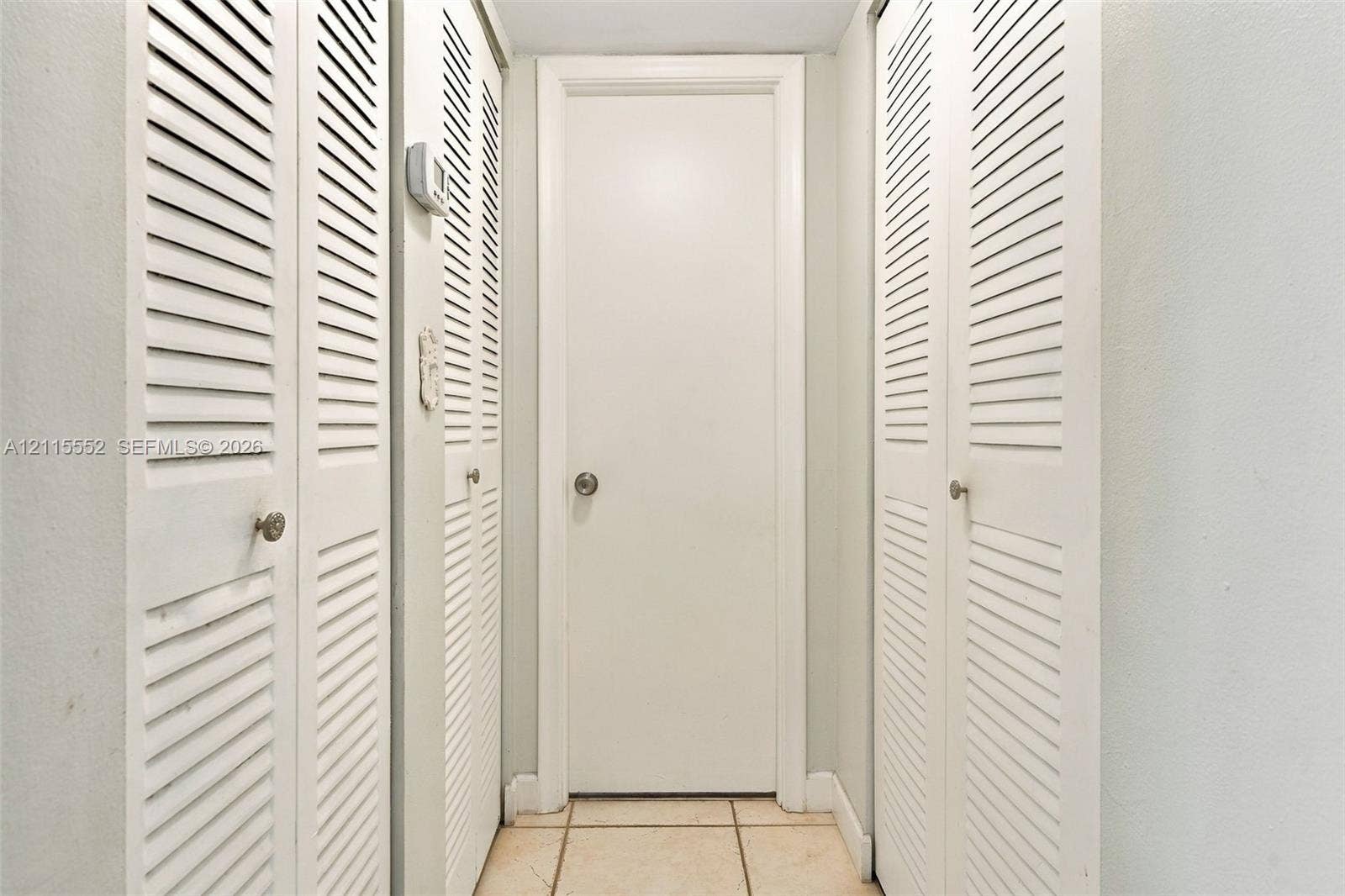 7805 Camino Real, #H-220, Miami, FL 33143 Hallway featuring dual sets of white louvered doors, a solid-panel door with a metallic knob, and light-toned tile flooring