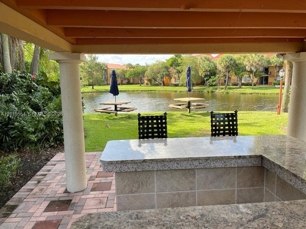 10781 Cleary Boulevard, #210, Plantation, Plantation, FL 33324 Covered outdoor kitchen featuring a stone-tile facade, stone countertop, and brick paver flooring, situated under a timber-framed pergola with structural columns overlooking a pond