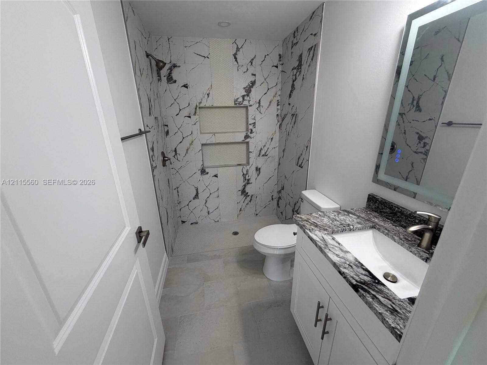 105 Concord Loop, Labelle, FL 33935 Bathroom featuring a stone-surface vanity with an integrated sink, illuminated vanity mirror, walk-in shower with dual recessed niches, and large-format tile flooring