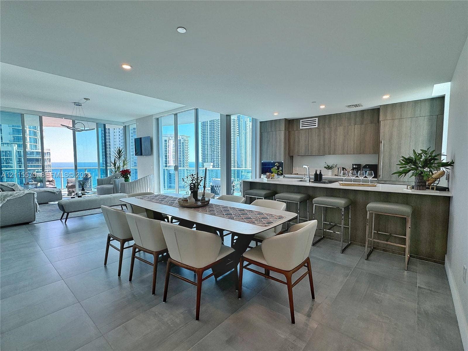 17550 Collins Avenue, #1401, Sunny Isles Beach, Sunny Isles Beach, FL 33160 Open-concept living space featuring expansive floor-to-ceiling glass doors, wood-finish cabinetry, a large kitchen island with integrated seating, and neutral stone-finish flooring