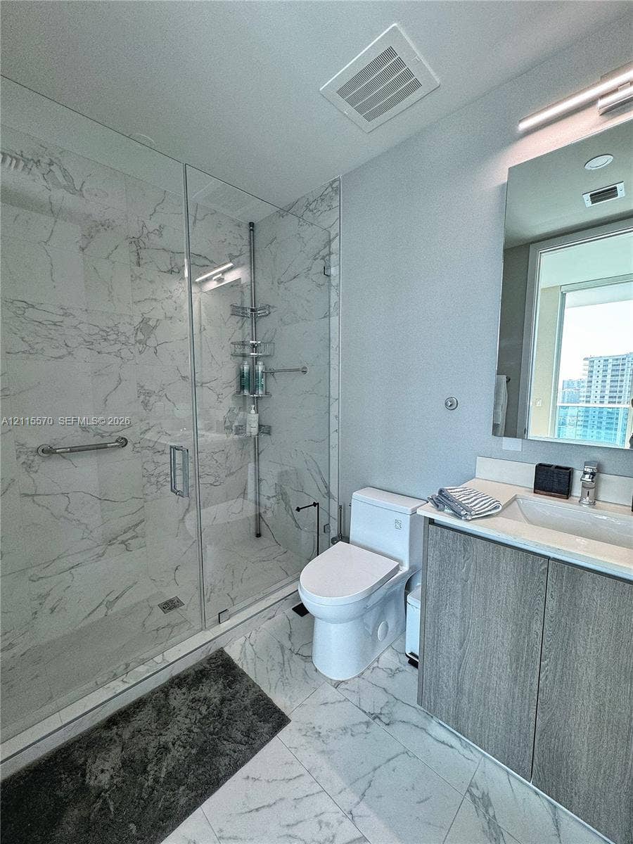 17550 Collins Avenue, #1401, Sunny Isles Beach, Sunny Isles Beach, FL 33160 Bathroom featuring a glass-enclosed walk-in shower with floor-to-ceiling stone-finish tile, vanity with integrated basin and wood-finish cabinetry, and oversized wall mirror