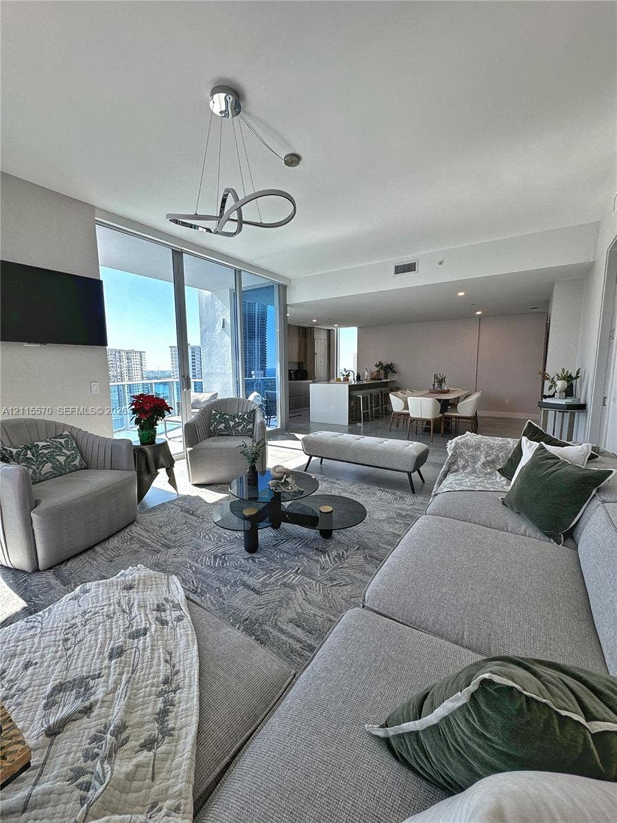 17550 Collins Avenue, #1401, Sunny Isles Beach, Sunny Isles Beach, FL 33160 Open-concept living space featuring floor-to-ceiling glass sliding doors, modern brushed-metal sculptural chandelier, kitchen island with stone countertop, and neutral-toned flooring