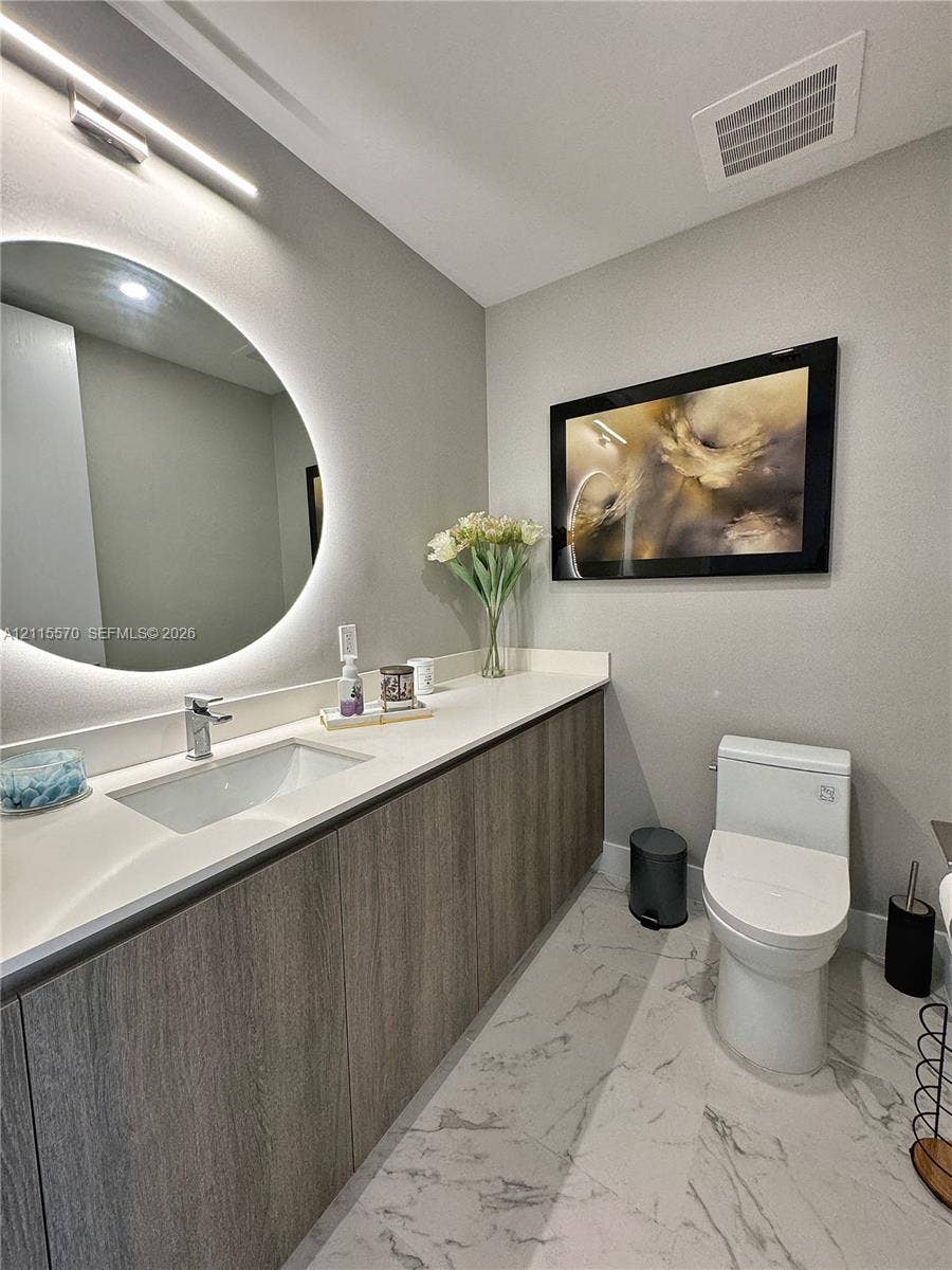 17550 Collins Avenue, #1401, Sunny Isles Beach, Sunny Isles Beach, FL 33160 Modern bathroom featuring a wood-finish floating vanity, integrated stone countertop with an undermount sink, LED-backlit circular mirror, and large-format veined tile flooring