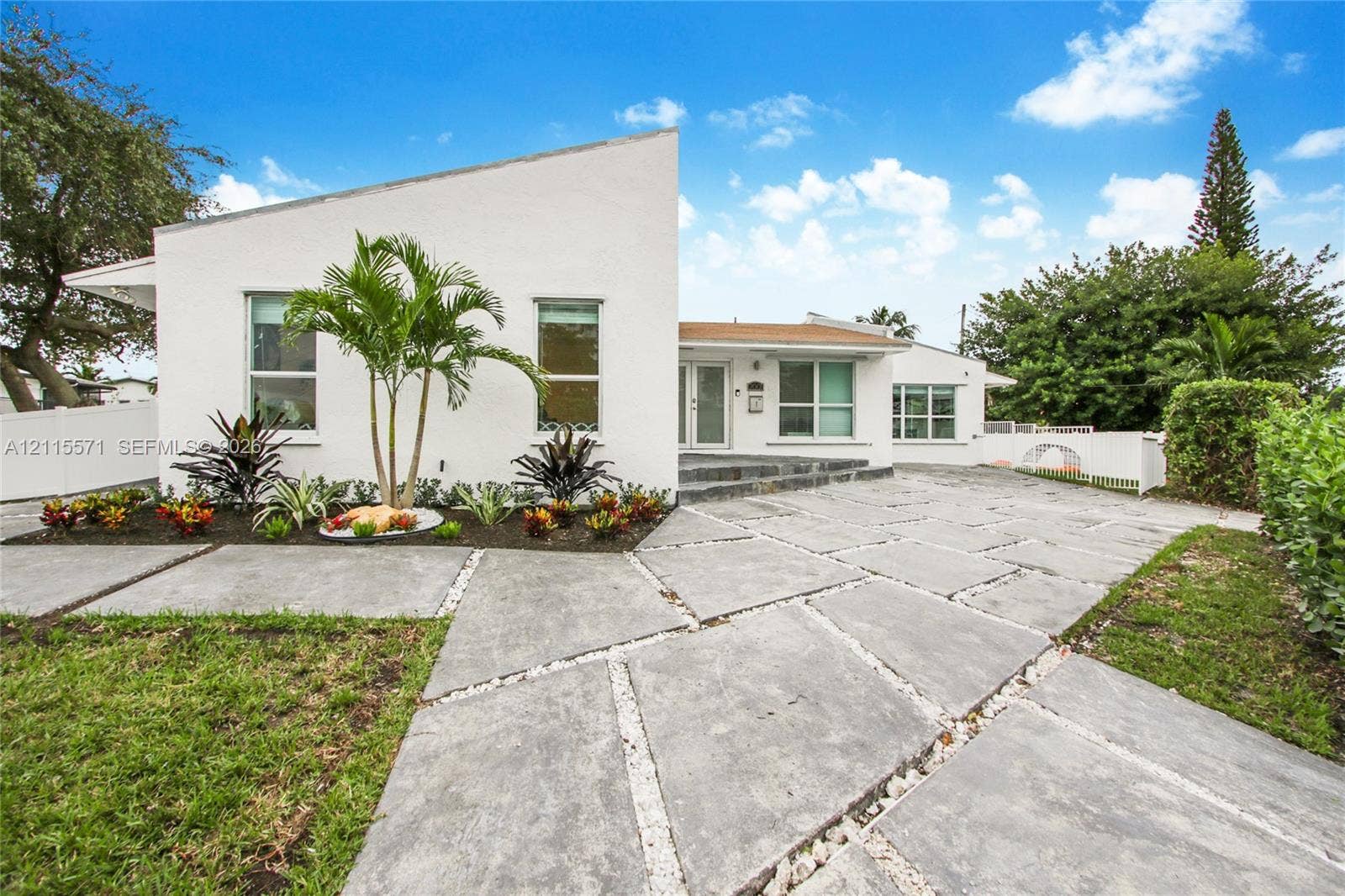 2001 NE 122nd Road, North Miami, FL 33181 Modern architectural exterior featuring a geometric roofline, white stucco facade, large-format concrete paver driveway, integrated stone-accented entryway, and landscaped garden beds