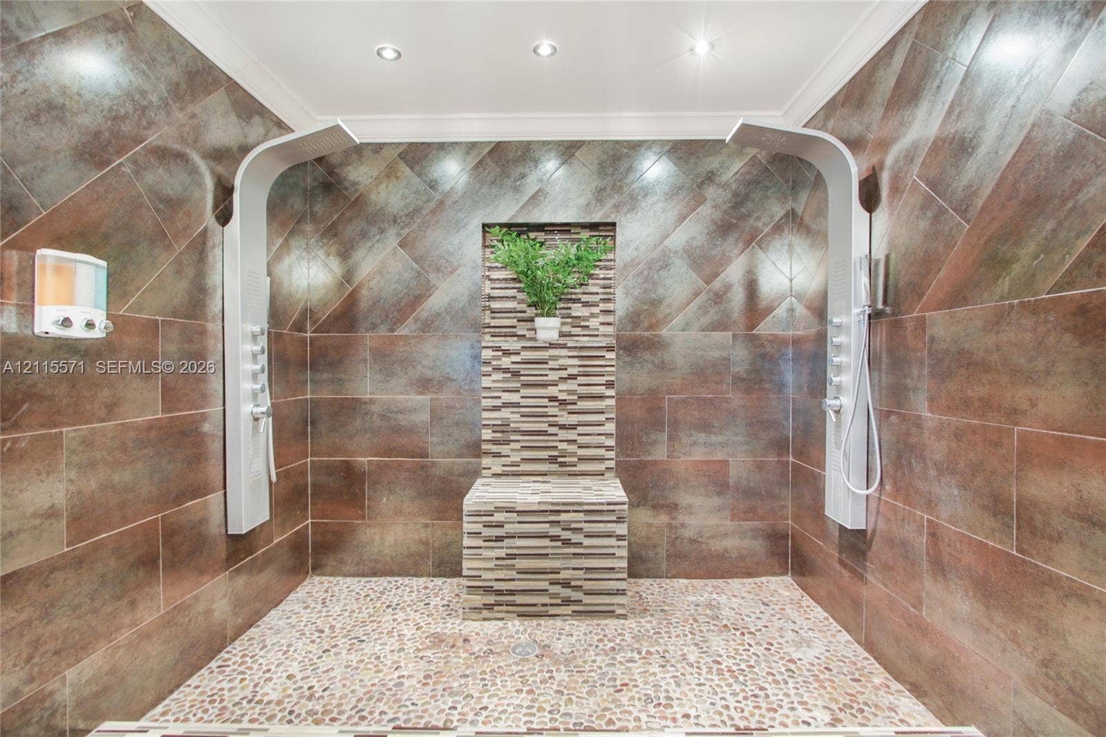2001 NE 122nd Road, North Miami, FL 33181 Walk-in shower featuring large-format metallic-finish wall tiles, dual vertical shower panels with integrated body sprayers, a central stone-tiled bench, and natural pebble flooring