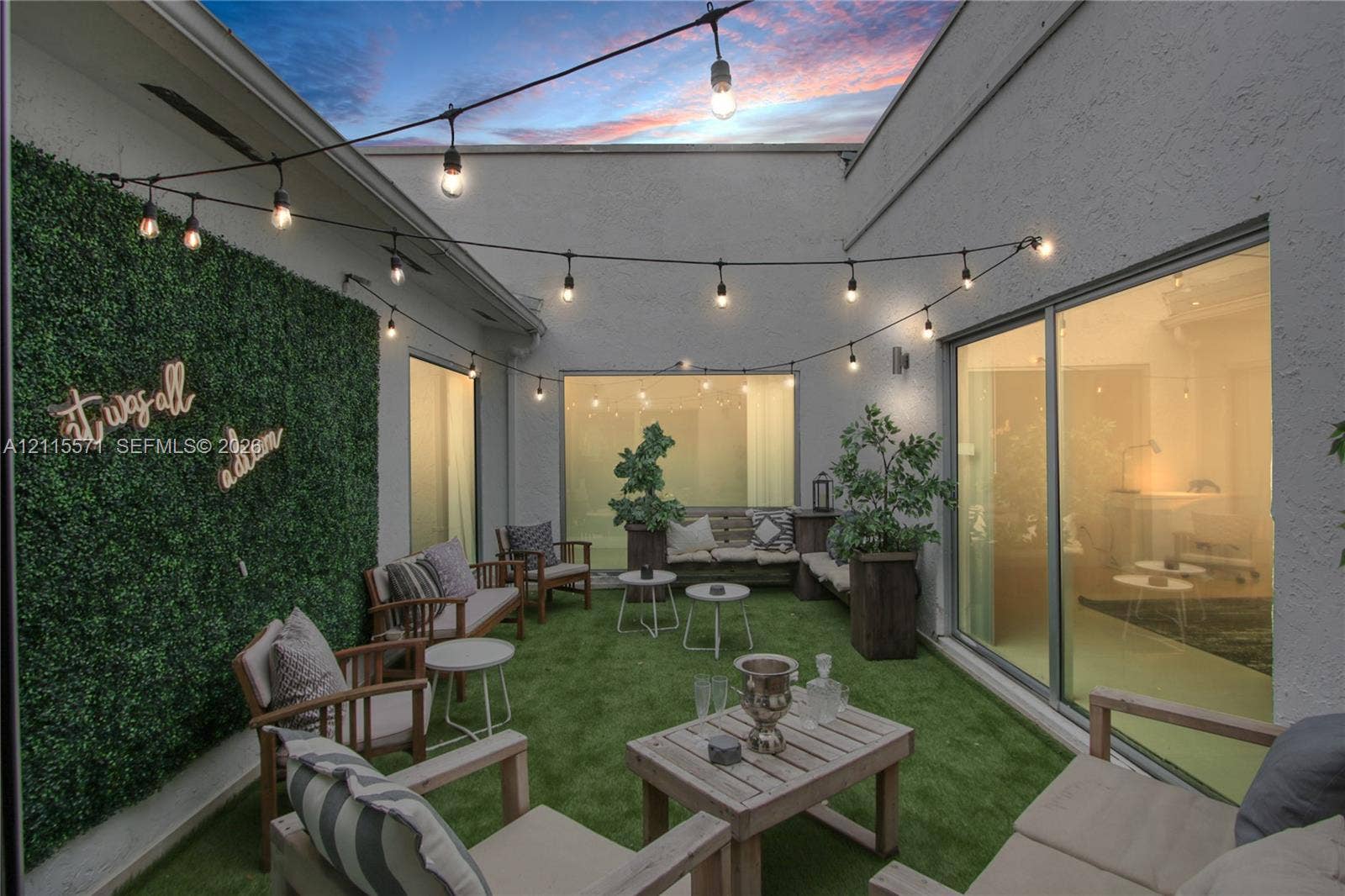 2001 NE 122nd Road, North Miami, FL 33181 Private courtyard featuring stucco walls, synthetic turf, integrated artificial greenery panel, and overhead string lighting, enclosed by floor-to-ceiling glass paneling