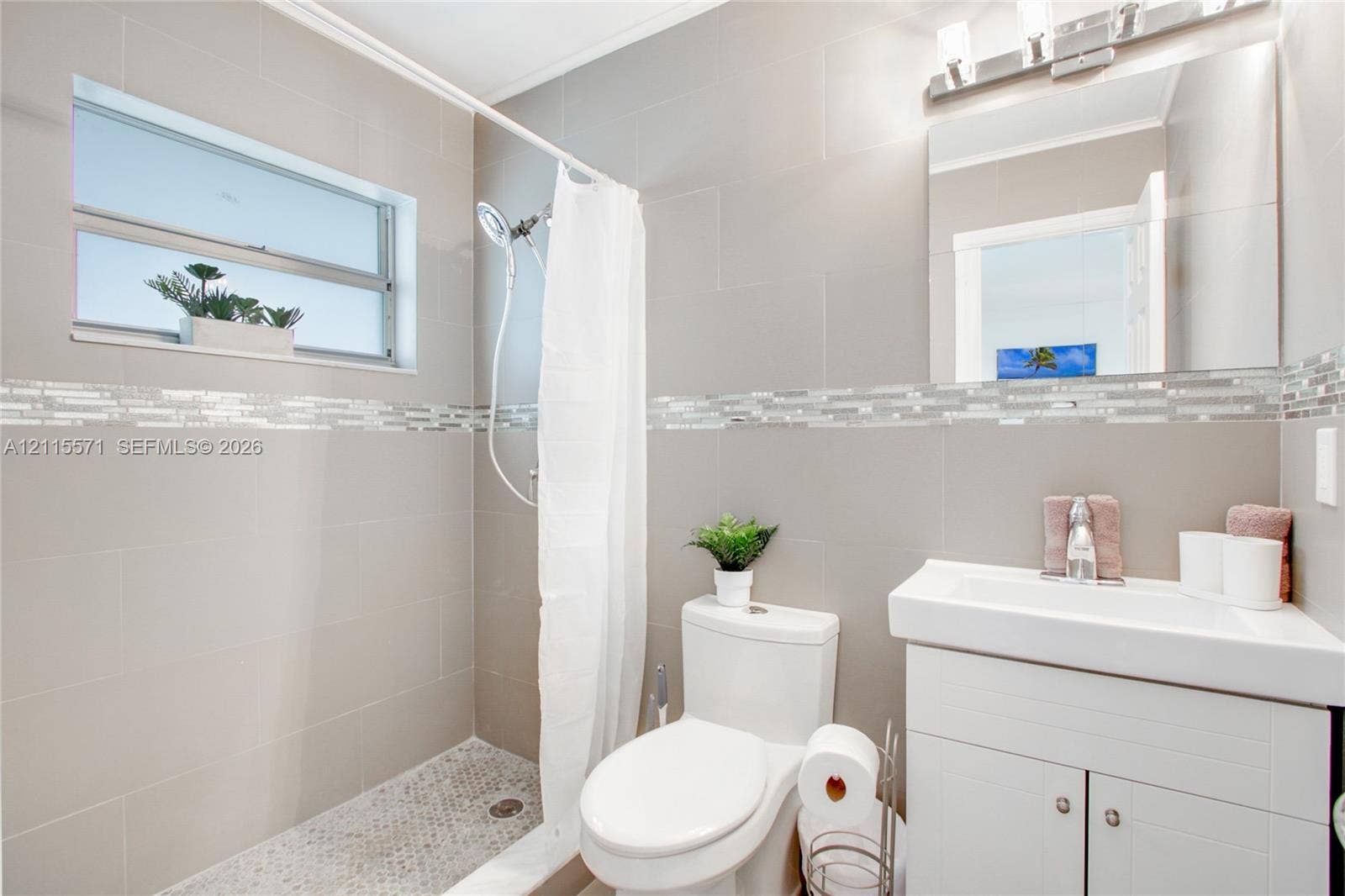 2001 NE 122nd Road, North Miami, FL 33181 Bathroom featuring large-format wall tile, glass mosaic accent border, pebble-style shower floor tiling, vanity with an integrated sink, and an expansive wall-mounted mirror