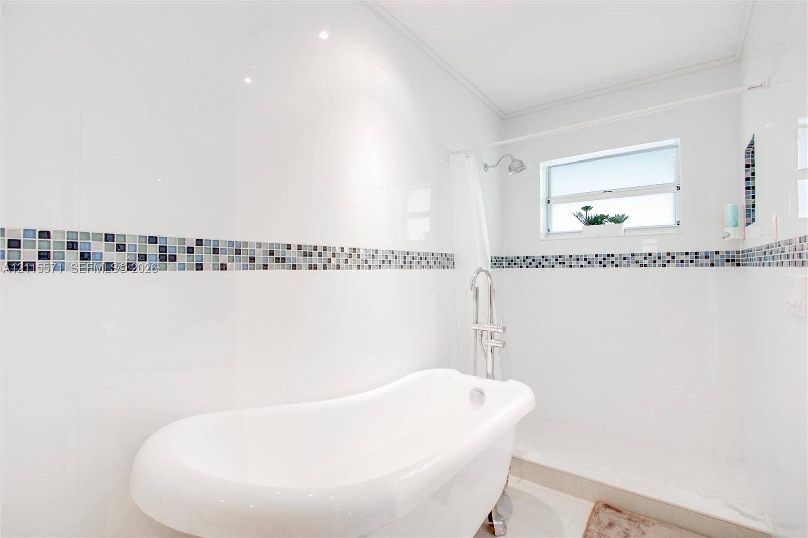 2001 NE 122nd Road, North Miami, FL 33181 Freestanding soaking tub with a chrome floor-mounted faucet, crisp white wall tile, decorative multi-tonal glass tile inlay, and recessed ceiling lighting