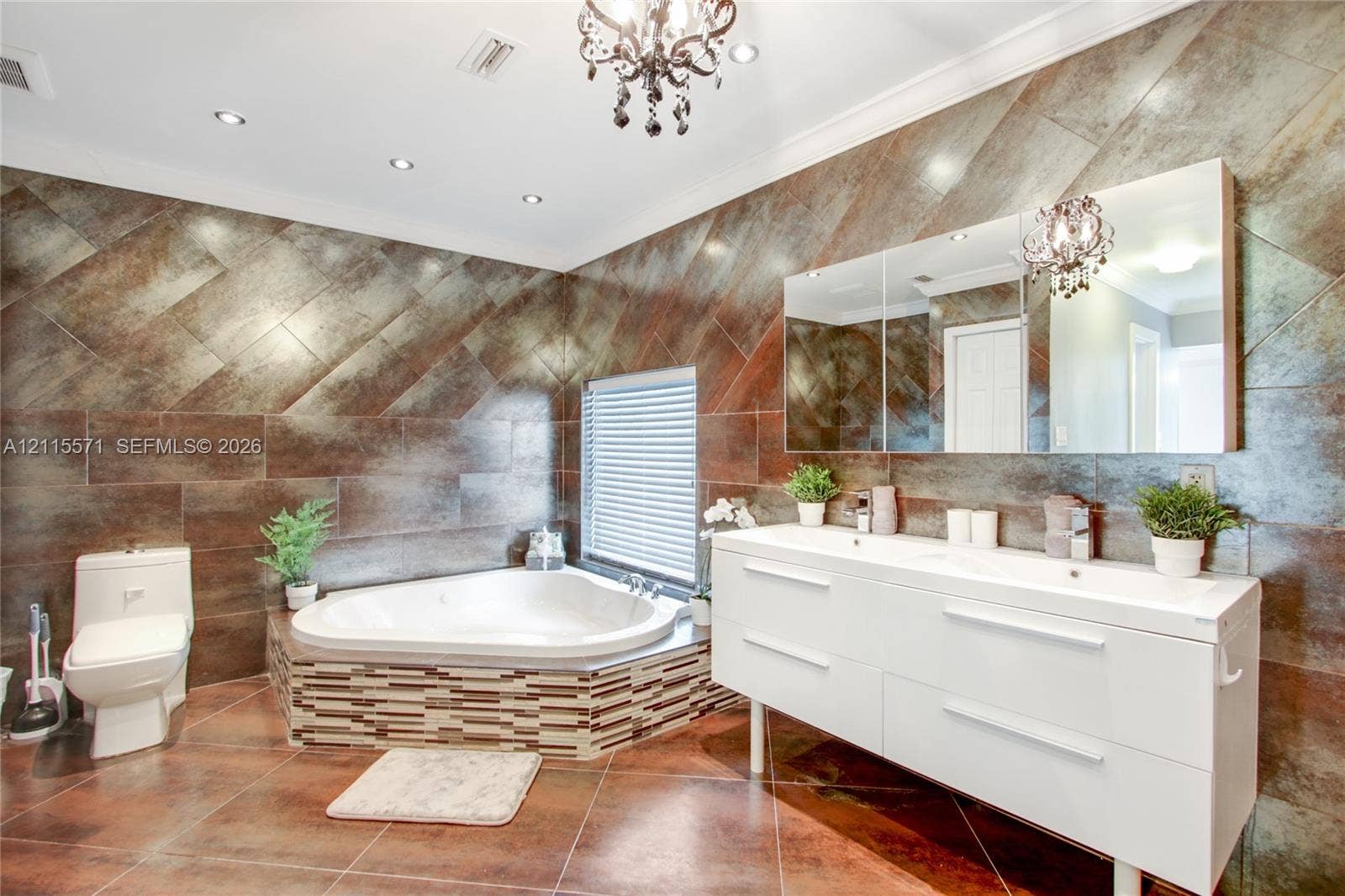 2001 NE 122nd Road, North Miami, FL 33181 Corner soaking tub with tiled surround, dual-sink white floating vanity, expansive wall-to-wall mirror, metallic-finish wall tiling, and decorative chandelier lighting