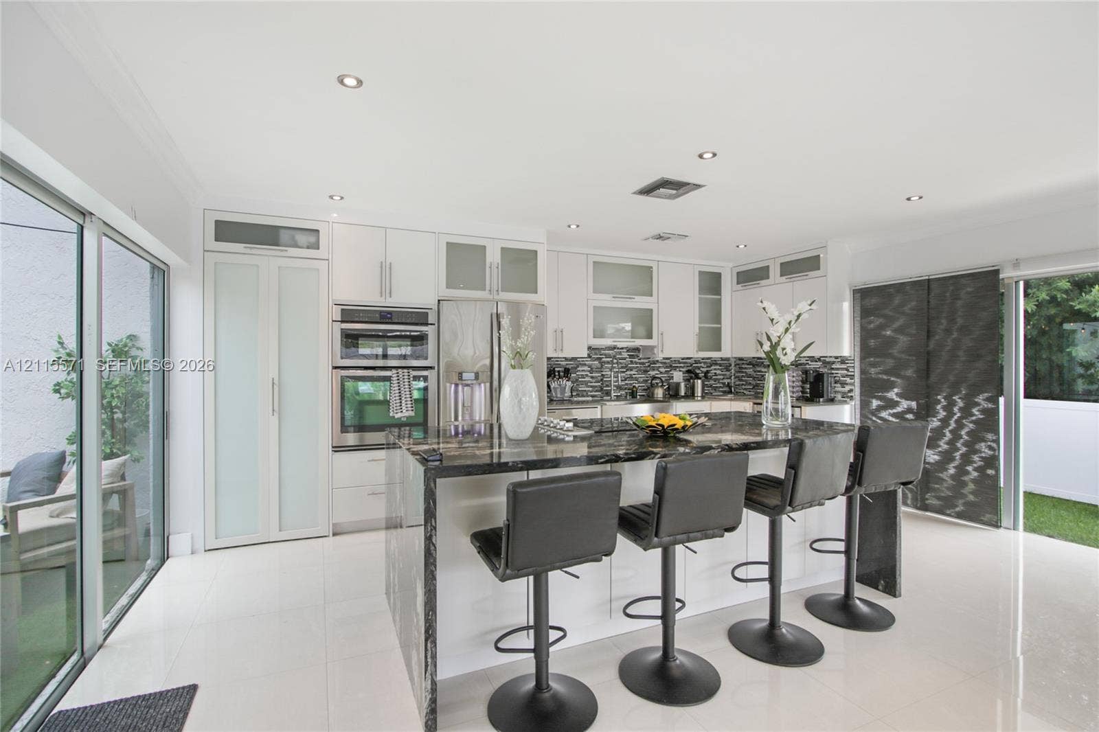 2001 NE 122nd Road, North Miami, FL 33181 Kitchen featuring white cabinetry with glass-panel accents, stone-topped central island, built-in stainless steel double ovens, patterned tile backsplash, and polished tile flooring