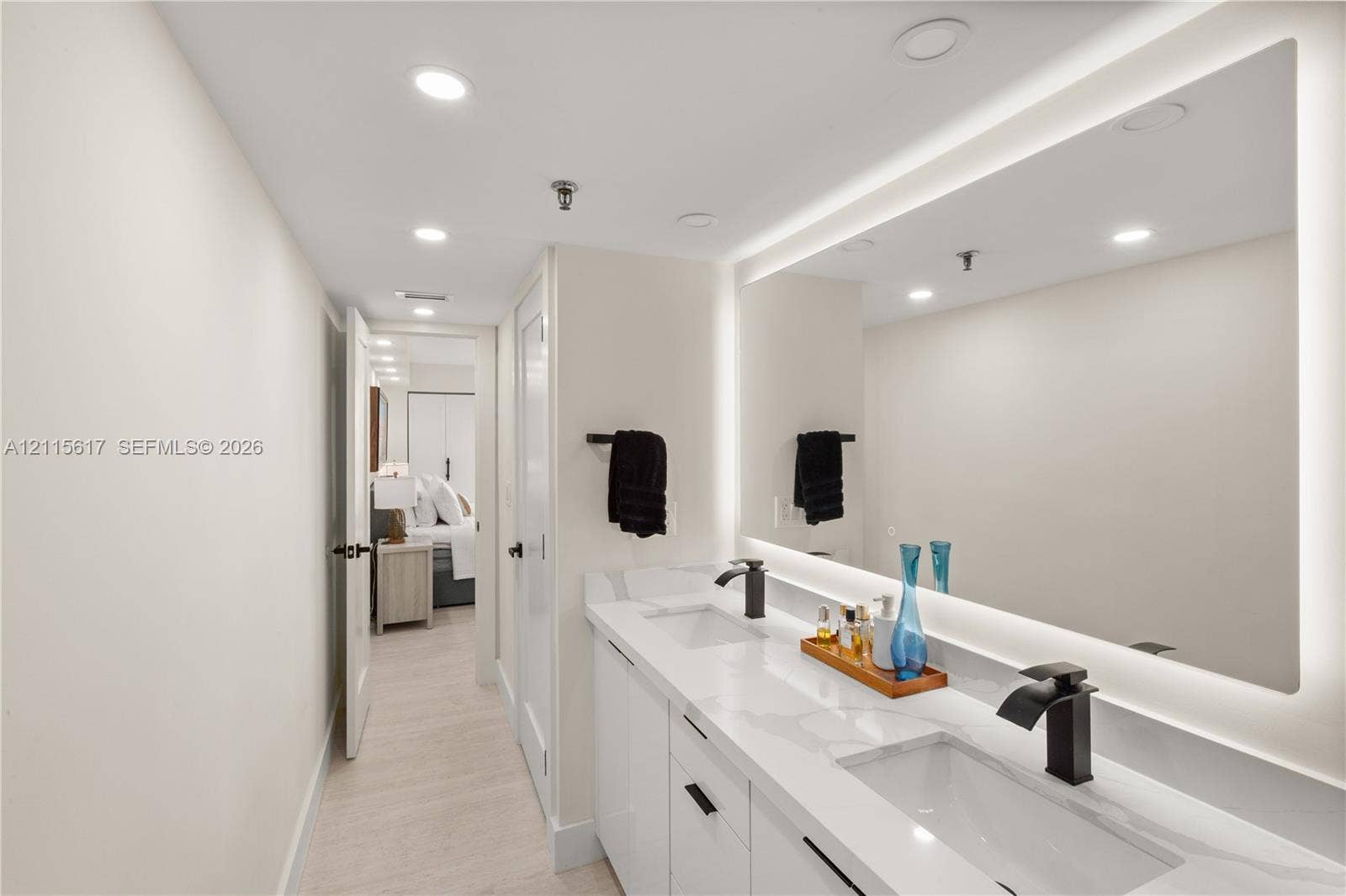 Double vanity featuring a light-toned countertop, integrated rectangular sinks, matte black widespread faucets, and a large backlit mirror
