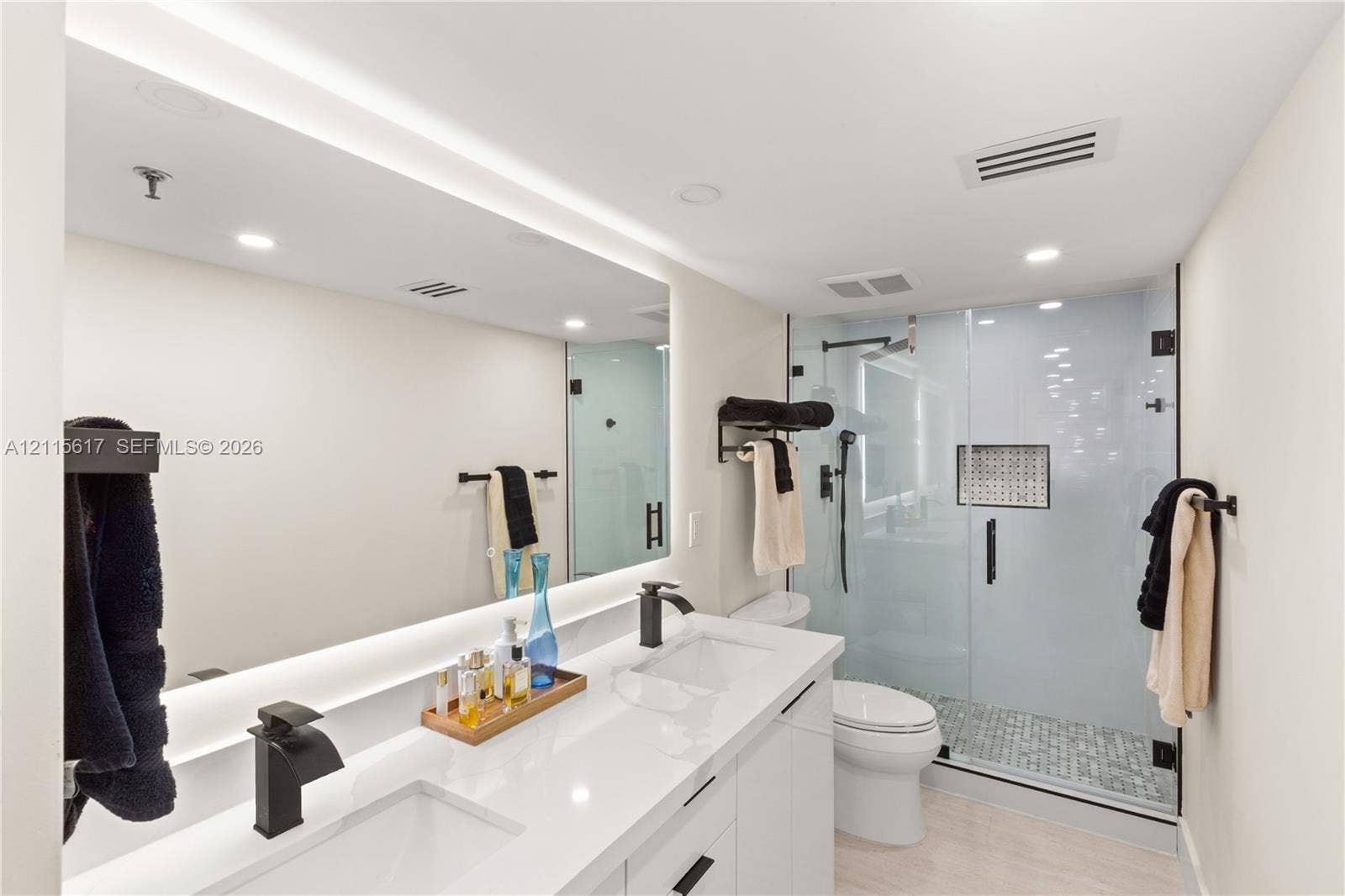 Dual-sink vanity with white stone-finish countertop, backlit mirror, matte black fixtures, and walk-in shower with glass enclosure