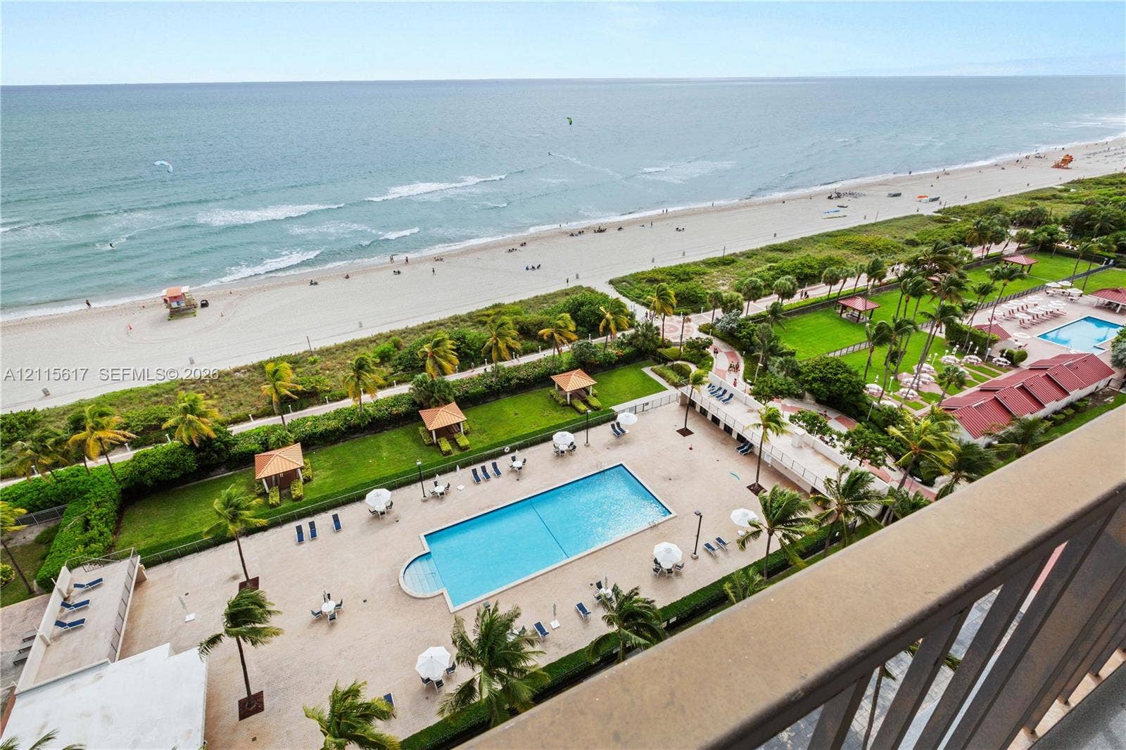 Elevated balcony featuring a metal railing, overlooking a rectangular swimming pool, manicured lawn areas with shade structures, and direct oceanfront access