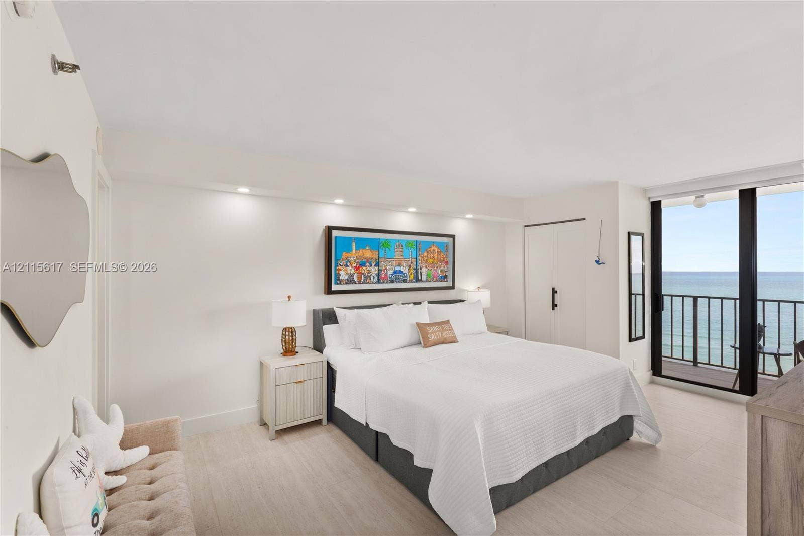 Neutral-toned interior space featuring light-colored flooring, recessed ceiling lighting, a built-in closet with matte black hardware, and floor-to-ceiling glass sliding doors opening to a balcony with an ocean backdrop