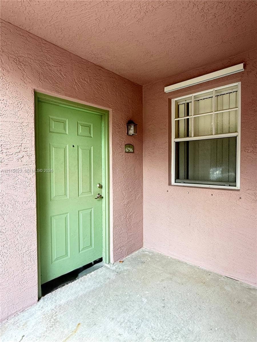 6796 W Sample Road, #6796, Coral Springs, Coral Springs, FL 33067 Covered entryway featuring a textured stucco facade, a six-panel entry door, a single-hung window with muntins, and a wall-mounted exterior light fixture