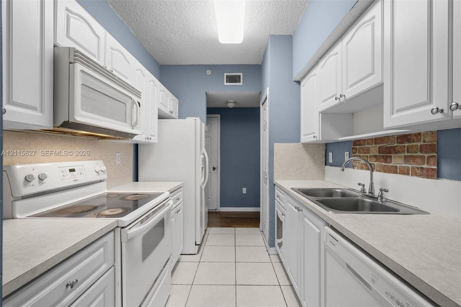 7085 Nova Drive, #218, Davie, FL 33317 Galley kitchen featuring white cabinetry, light-toned countertops, tile flooring, an exposed brick backsplash, and a double-basin stainless steel sink