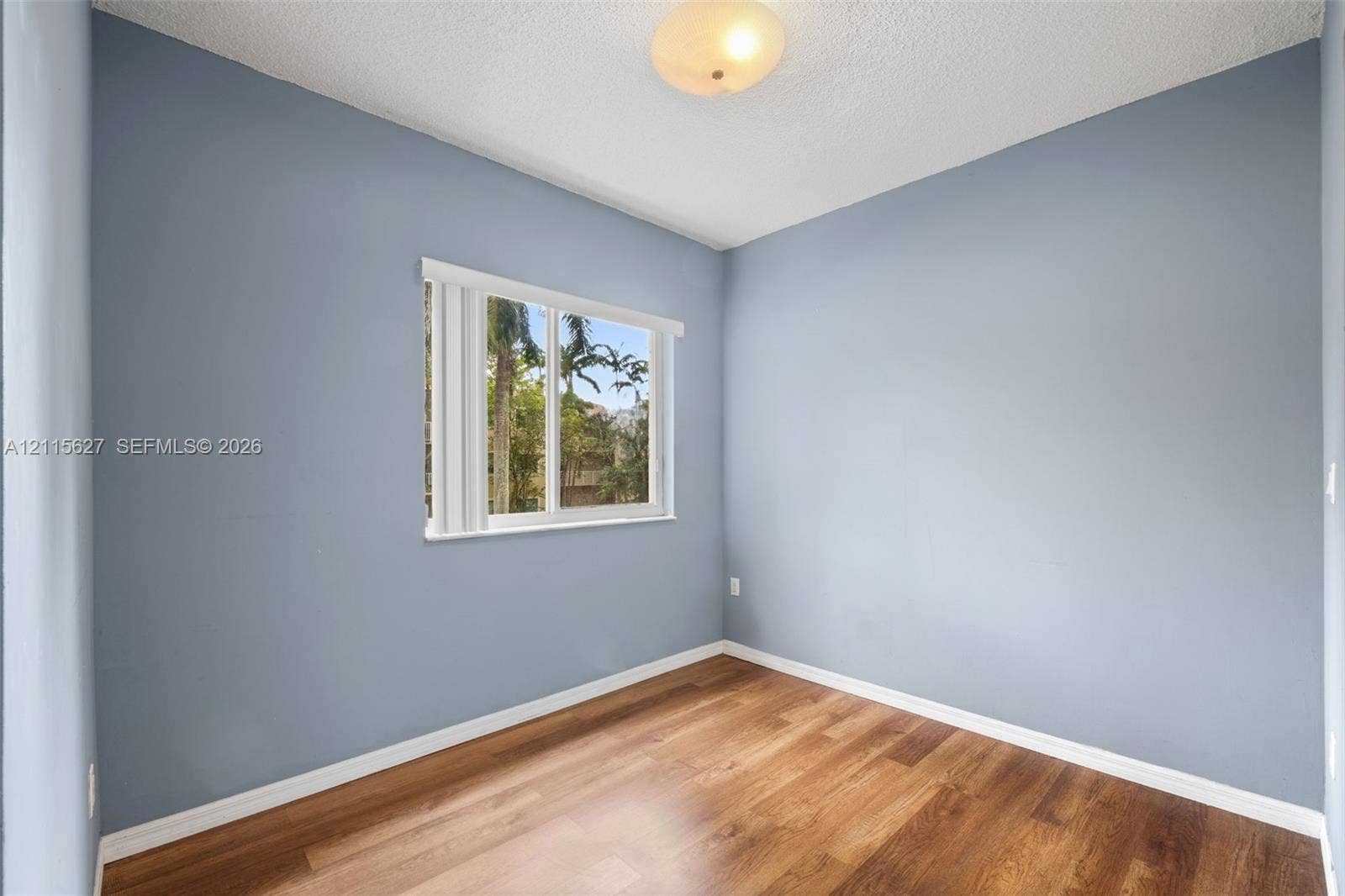 7085 Nova Drive, #218, Davie, FL 33317 Room featuring wood-finish flooring, neutral blue-toned wall paint, a single window with vertical blinds, and a central ceiling-mounted light fixture