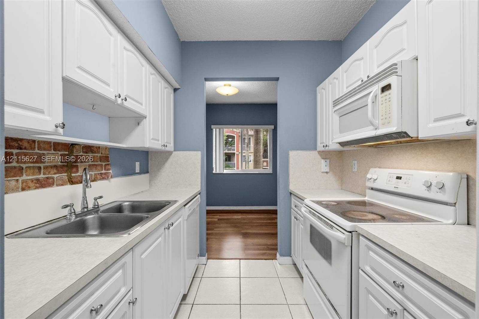 7085 Nova Drive, #218, Davie, FL 33317 Kitchen featuring white shaker-style cabinetry, a brick backsplash, dual-basin stainless steel sink, tile flooring, and integrated white appliances