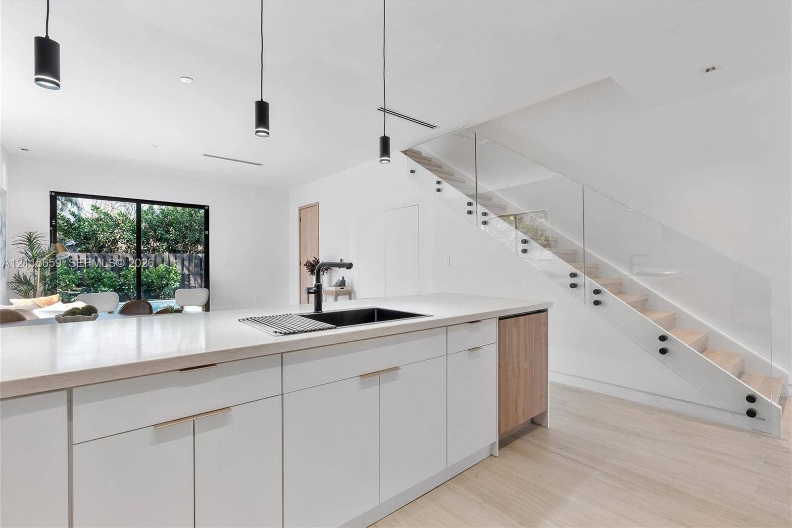 4719 N Miami Avenue, Miami, FL 33127 Modern kitchen island featuring a quartz countertop, black undermount sink, and matte black faucet, complemented by white cabinetry, wood-finish flooring, and a glass-paneled staircase