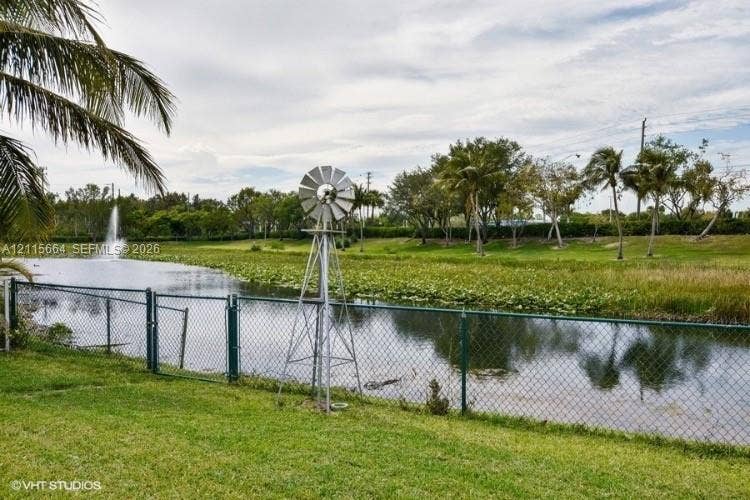 19409 NW 24th Place, Pembroke Pines, FL 33029 Waterfront parcel featuring a pond with a fountain, a metal windmill, palm trees, and perimeter chain-link fencing